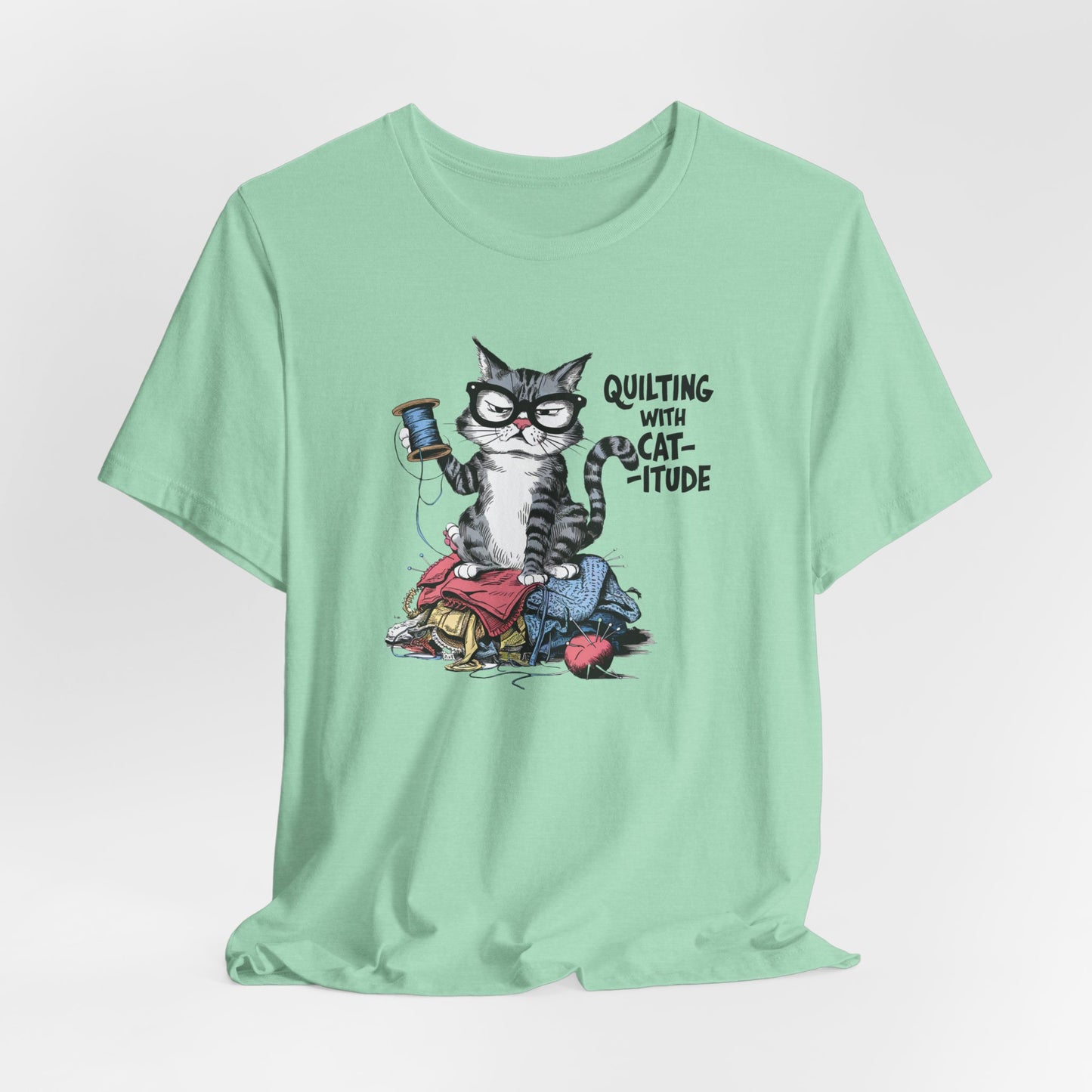 A Heather Mint t-shirt featuring a sassy cartoon cat sitting on a pile of colorful fabric, holding a spool of thread, with the phrase 'Quilting with Cat-itude,' perfect for quilters and cat lovers.