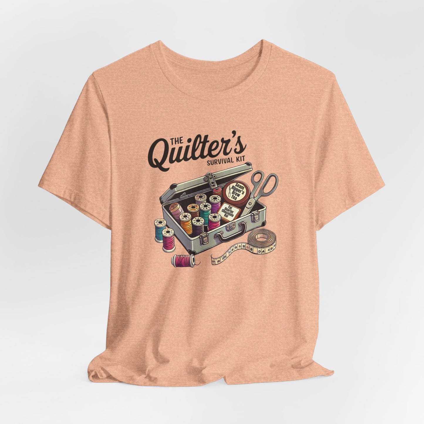 A Heather Peach t-shirt featuring a whimsical illustration of a quilter’s survival kit with colorful thread spools, scissors, and a tape measure, perfect for sewing and quilting enthusiasts.