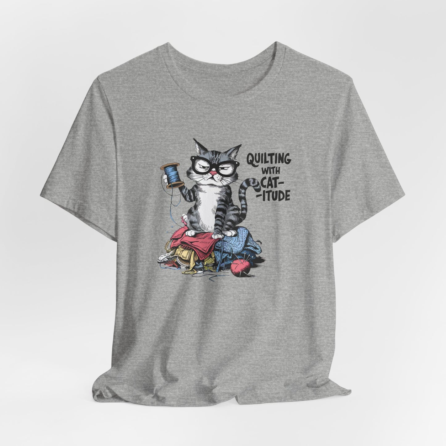 An Athletic Heather t-shirt featuring a sassy cartoon cat sitting on a pile of colorful fabric, holding a spool of thread, with the phrase 'Quilting with Cat-itude,' perfect for quilters and cat lovers.