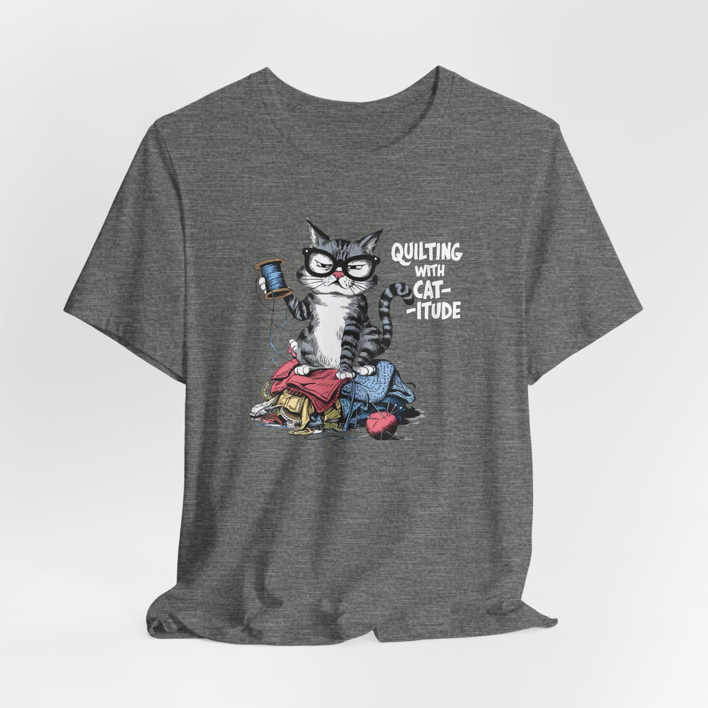 A Deep Heather t-shirt featuring a sassy cartoon cat sitting on a pile of colorful fabric, holding a spool of thread, with the phrase 'Quilting with Cat-itude,' perfect for quilters and cat lovers.