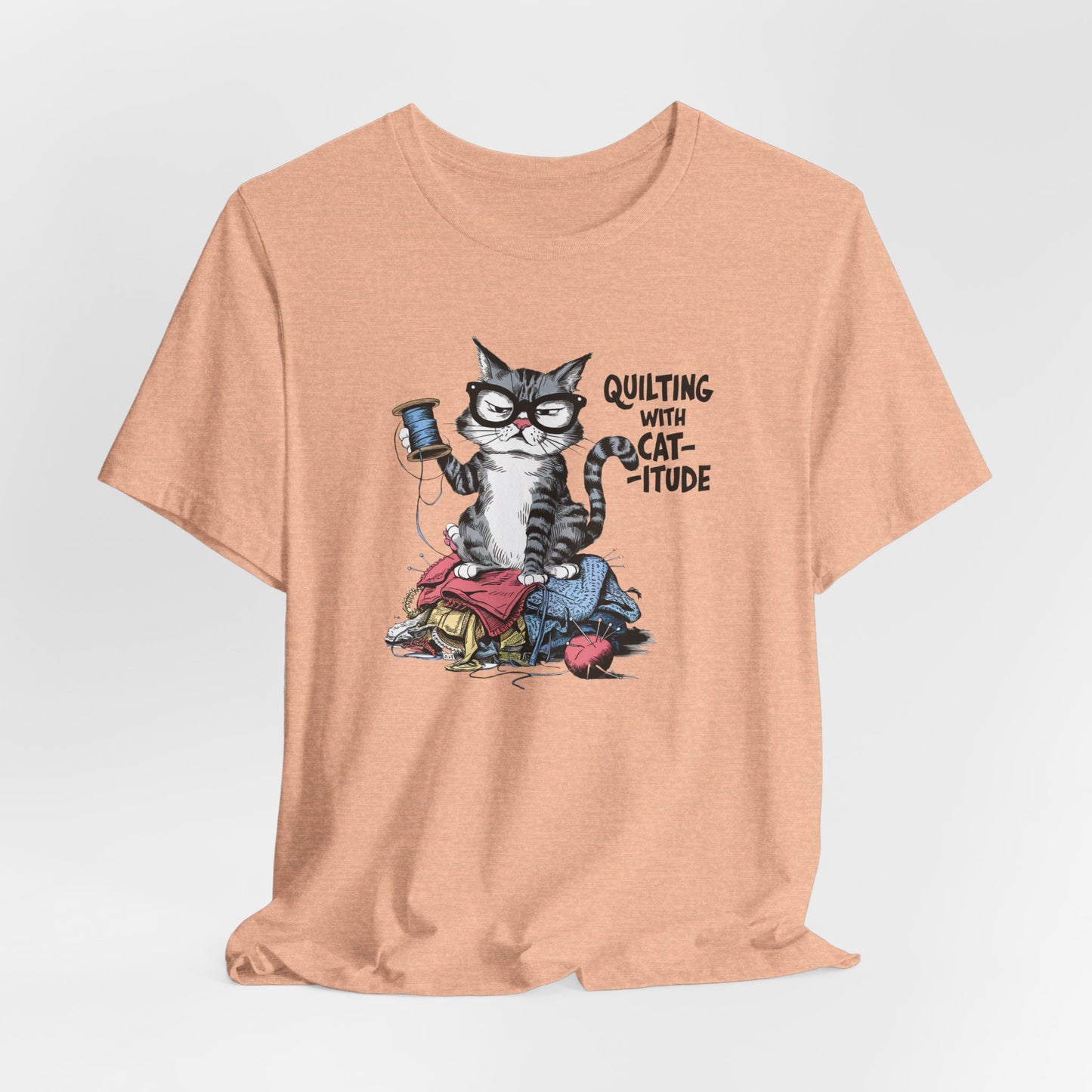 A Heather Peach t-shirt featuring a sassy cartoon cat sitting on a pile of colorful fabric, holding a spool of thread, with the phrase 'Quilting with Cat-itude,' perfect for quilters and cat lovers.