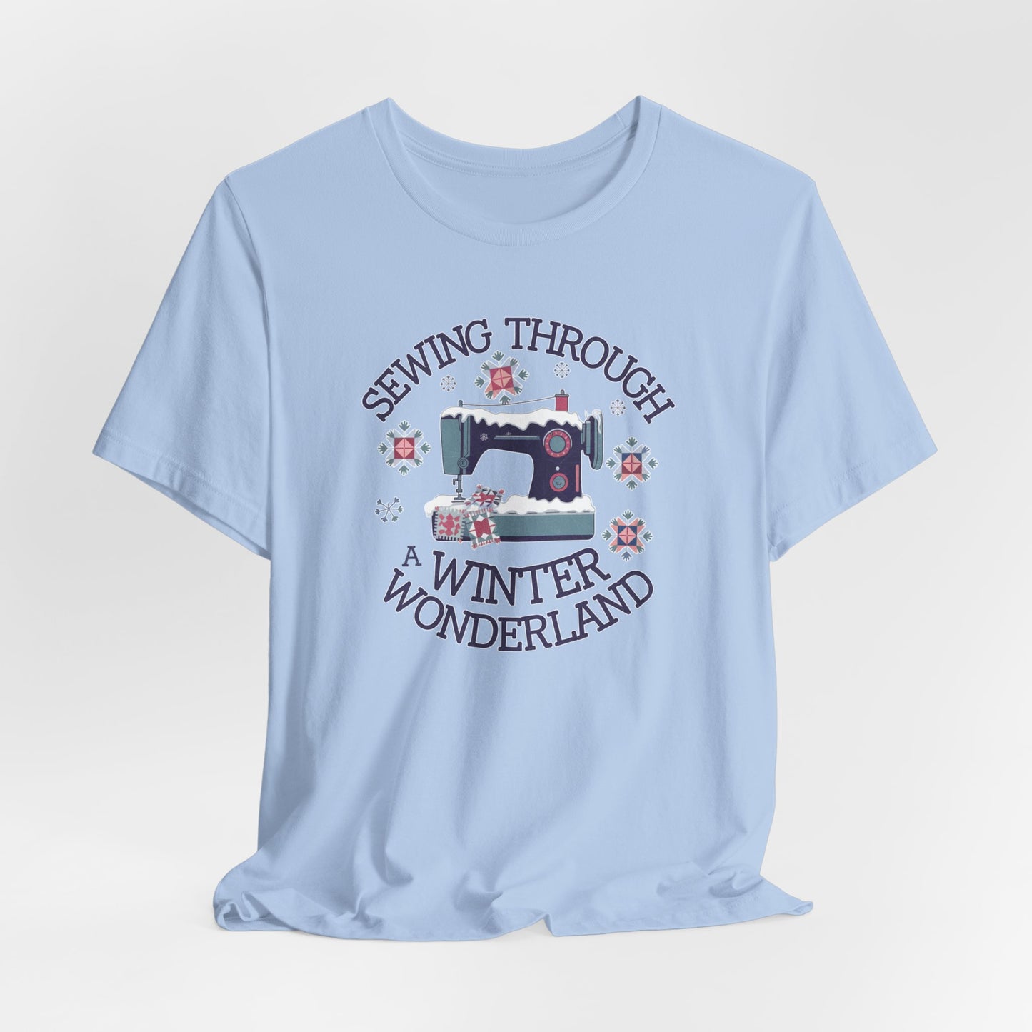 A Baby Blue winter quilting t-shirt featuring  a sewing machine covered in snow, surrounded by quilted snowflakes, with the phrase "Sewing Through a Winter Wonderland" above and below the image.