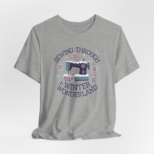 An Athletic Heather winter quilting t-shirt featuring  a sewing machine covered in snow, surrounded by quilted snowflakes, with the phrase "Sewing Through a Winter Wonderland" above and below the image.