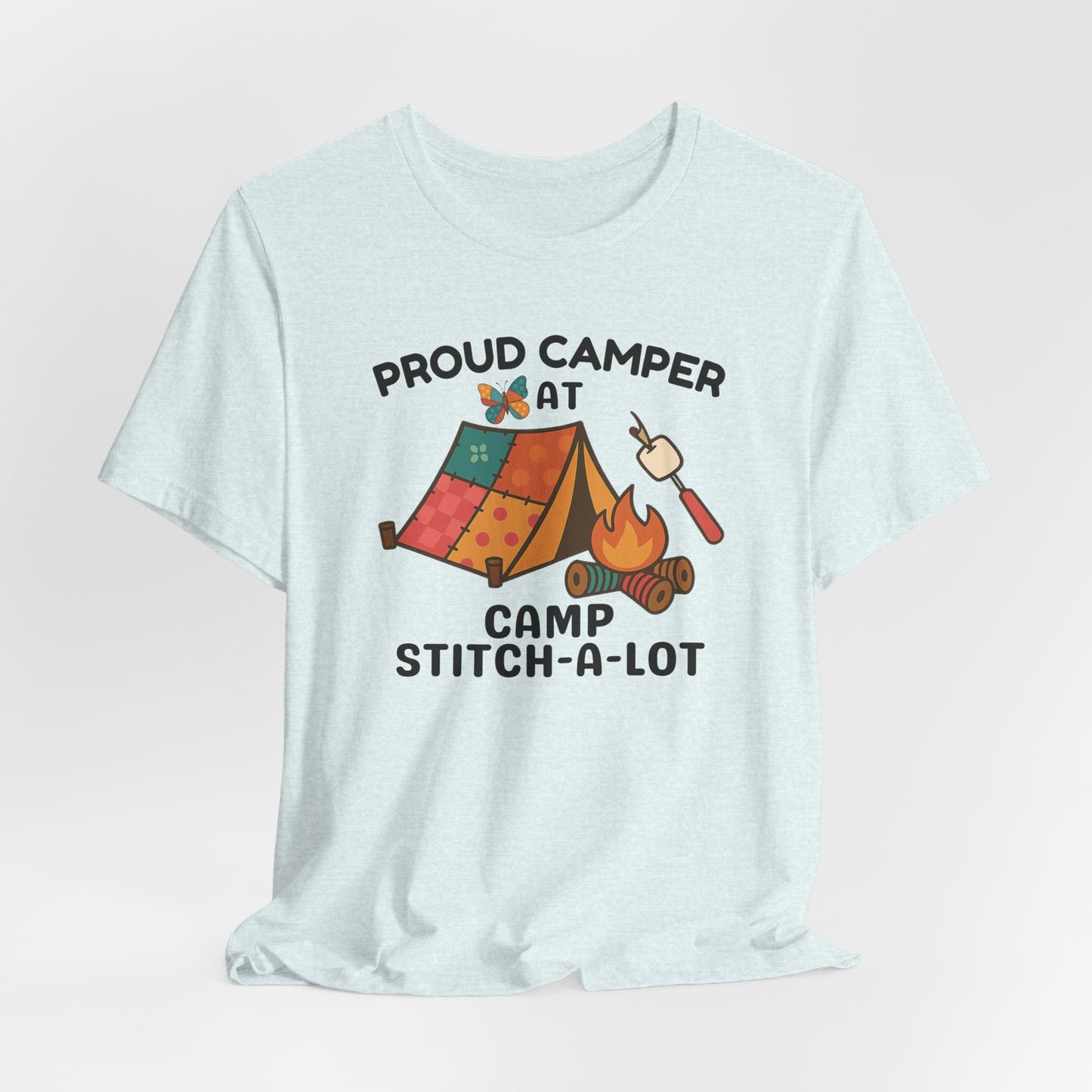 A A Heather Ice Blue t-shirt featuring a colorful design with a patchwork tent, thread spool campfire, marshmallow on a seam ripper stick, and a butterfly. Text reads "Proud Camper at Camp Stitch-a-Lot." Fun gift for quilting retreats.