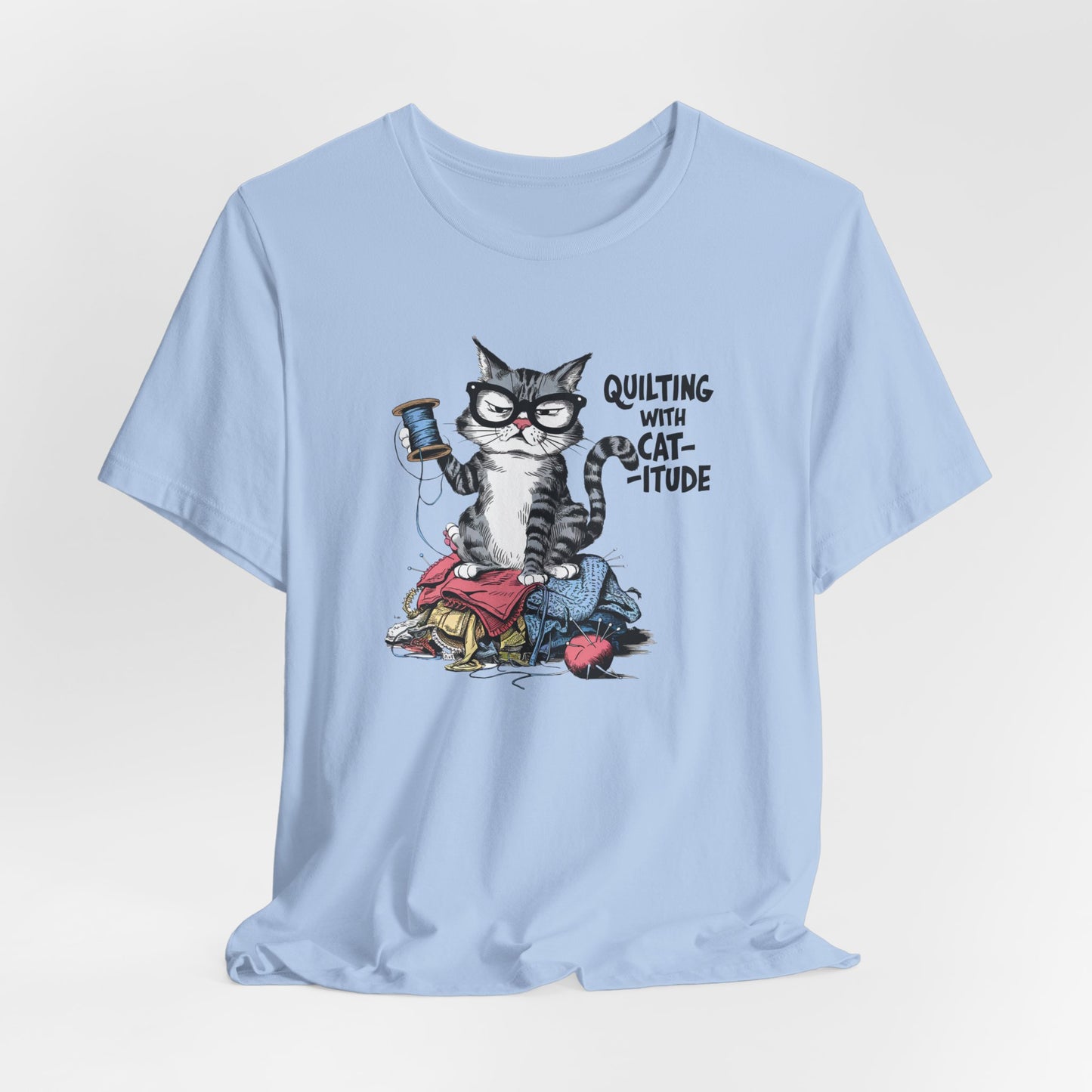 A Baby Blue t-shirt featuring a sassy cartoon cat sitting on a pile of colorful fabric, holding a spool of thread, with the phrase 'Quilting with Cat-itude,' perfect for quilters and cat lovers.