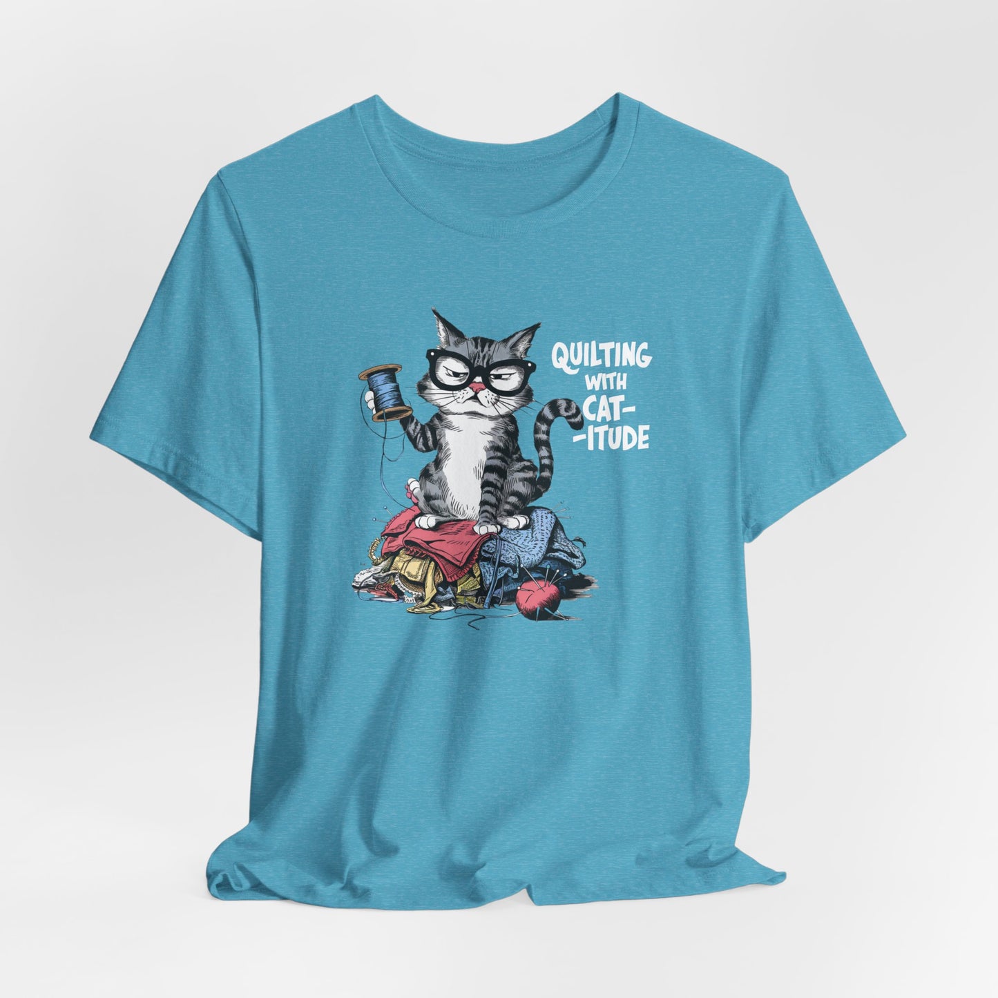 A Heather Aqua t-shirt featuring a sassy cartoon cat sitting on a pile of colorful fabric, holding a spool of thread, with the phrase 'Quilting with Cat-itude,' perfect for quilters and cat lovers.