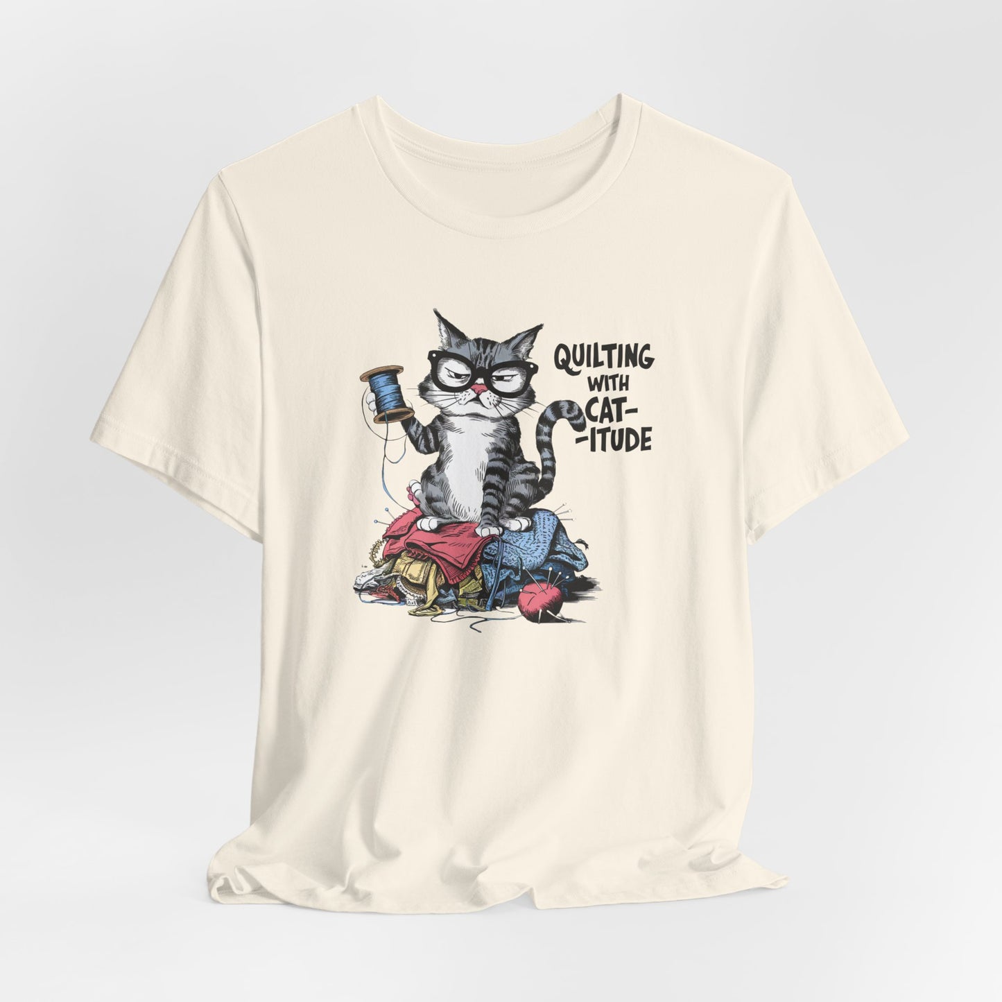 A Natural t-shirt featuring a sassy cartoon cat sitting on a pile of colorful fabric, holding a spool of thread, with the phrase 'Quilting with Cat-itude,' perfect for quilters and cat lovers.