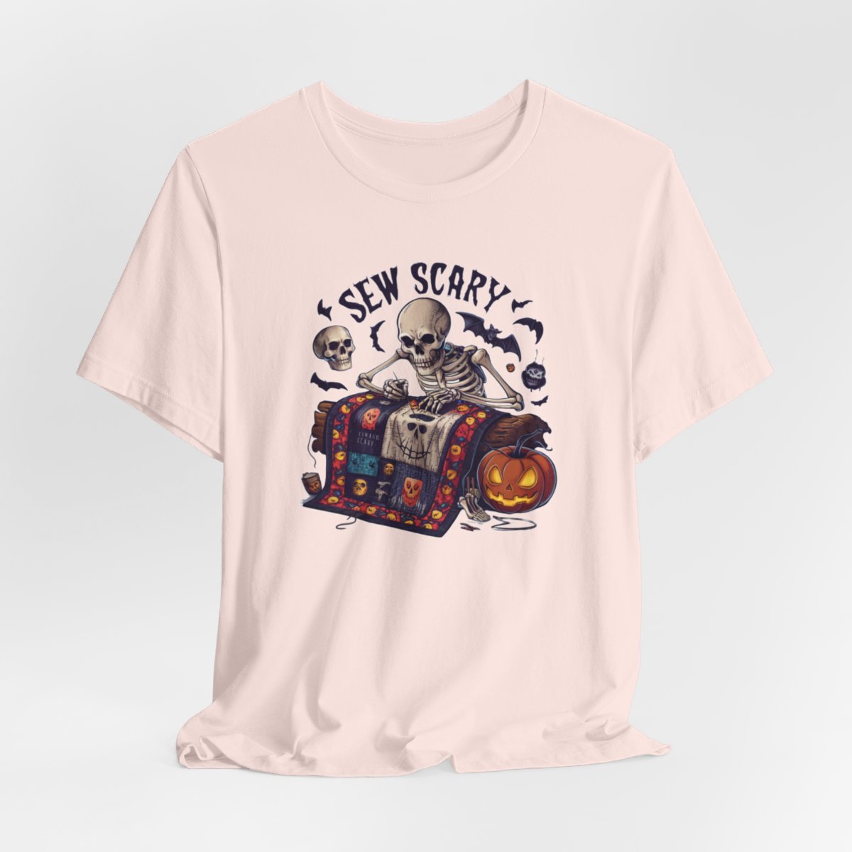 A Soft Pink Halloween Quilting T-Shirt featuring a skeleton sewing Halloween quilt. Colorful patchwork with spooky motifs. Bats and pumpkins surround skeleton. Text reads "Sew Scary". Ideal for quilters who love Halloween.
