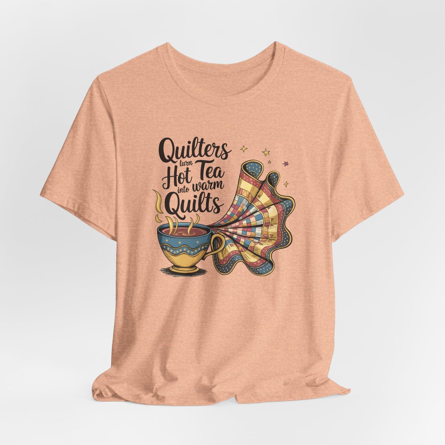 A Heather Peach t-shirt with a whimsical design of a teacup transforming into a patchwork quilt, accompanied by the phrase 'Quilters turn hot tea into warm quilts.' Ideal for quilters and tea lovers