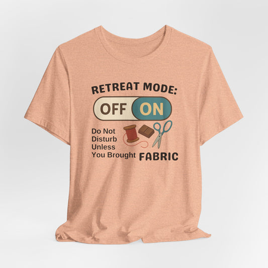A A Heather Peach t-shirt with a toggle switch that says "Retreat Mode: OFF/ON" surrounded by a spool of thread, chocolate, and scissors. Text below reads "Do not disturb unless you brought fabric." Fun gift for quilting retreats.