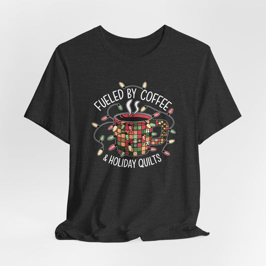 A Dark Grey Heather christmas quilting t-shirt featuring a cozy coffee mug covered in patchwork quilt patterns, wrapped in festive holiday lights, with the phrase "Fueled by Coffee & Holiday Quilts" surrounding the design.