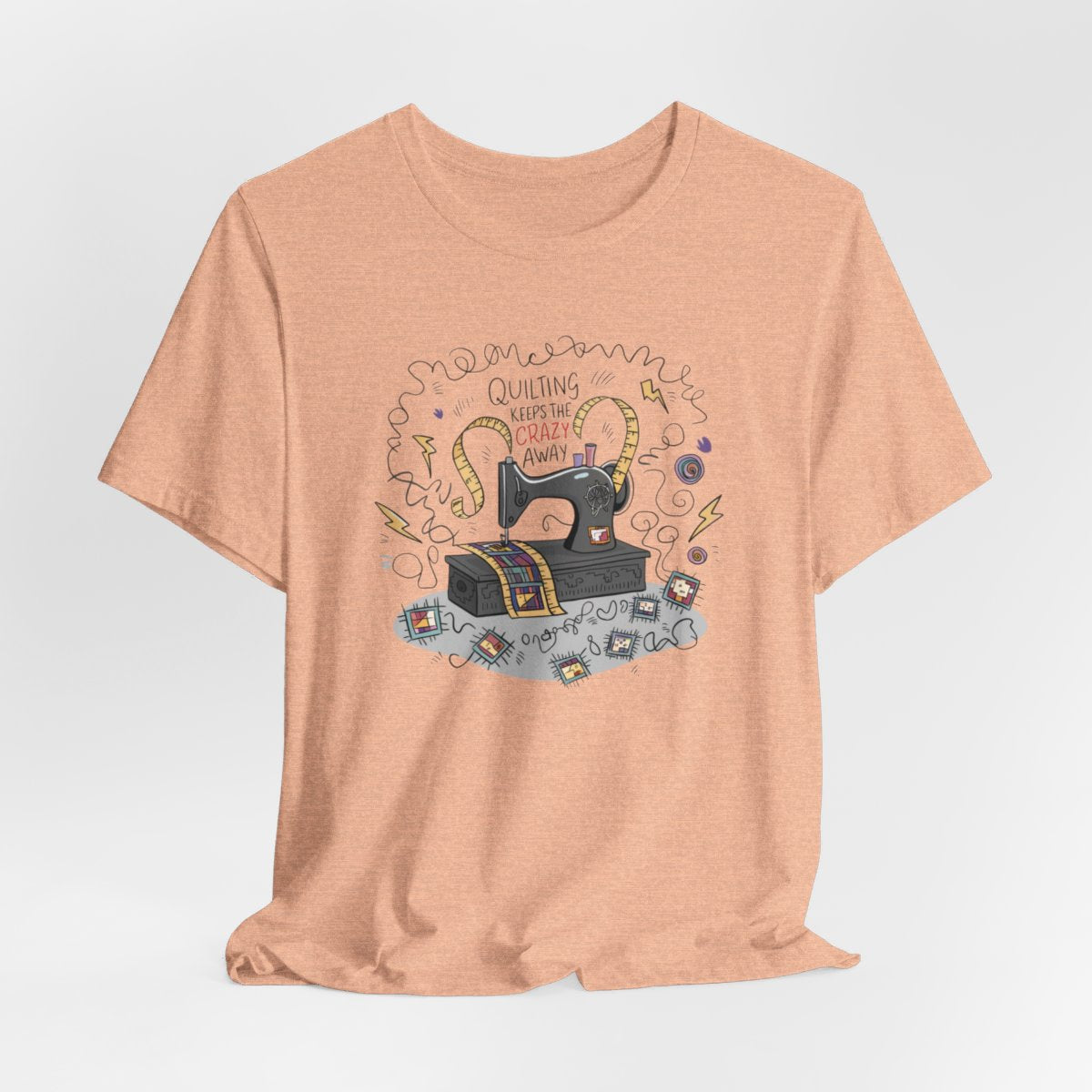 A Heather Peach T-Shirt with a vintage sewing machine, measuring tape heart, and text "Quilting Keeps the Crazy Away" amidst swirls and quilting motifs.