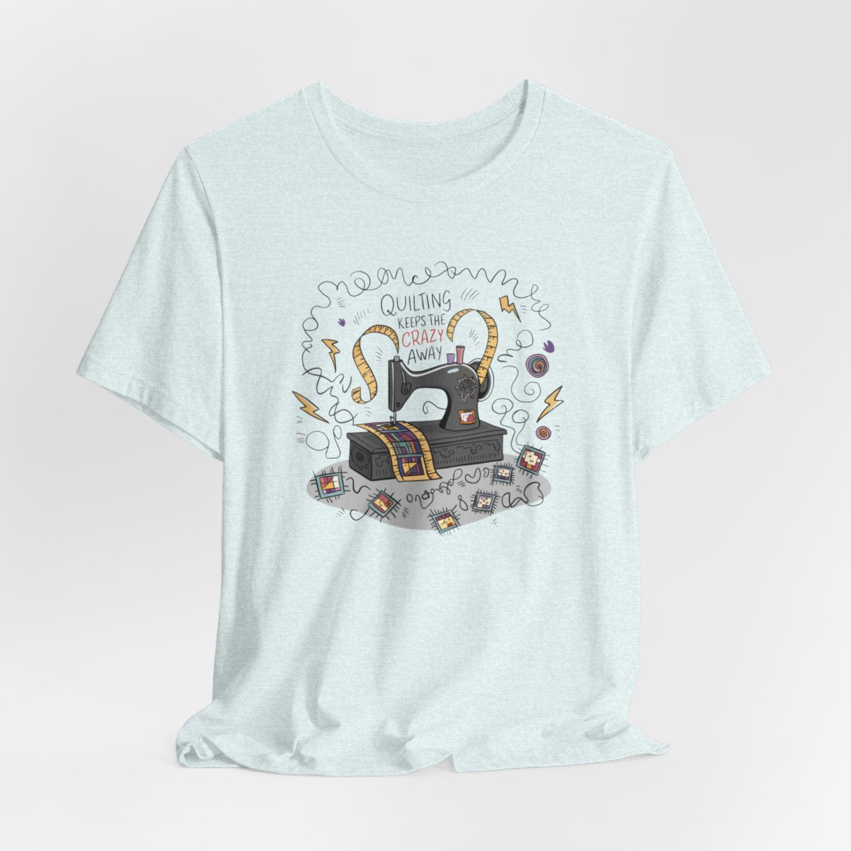 A Heather Ice Blue T-Shirt with a vintage sewing machine, measuring tape heart, and text "Quilting Keeps the Crazy Away" amidst swirls and quilting motifs.