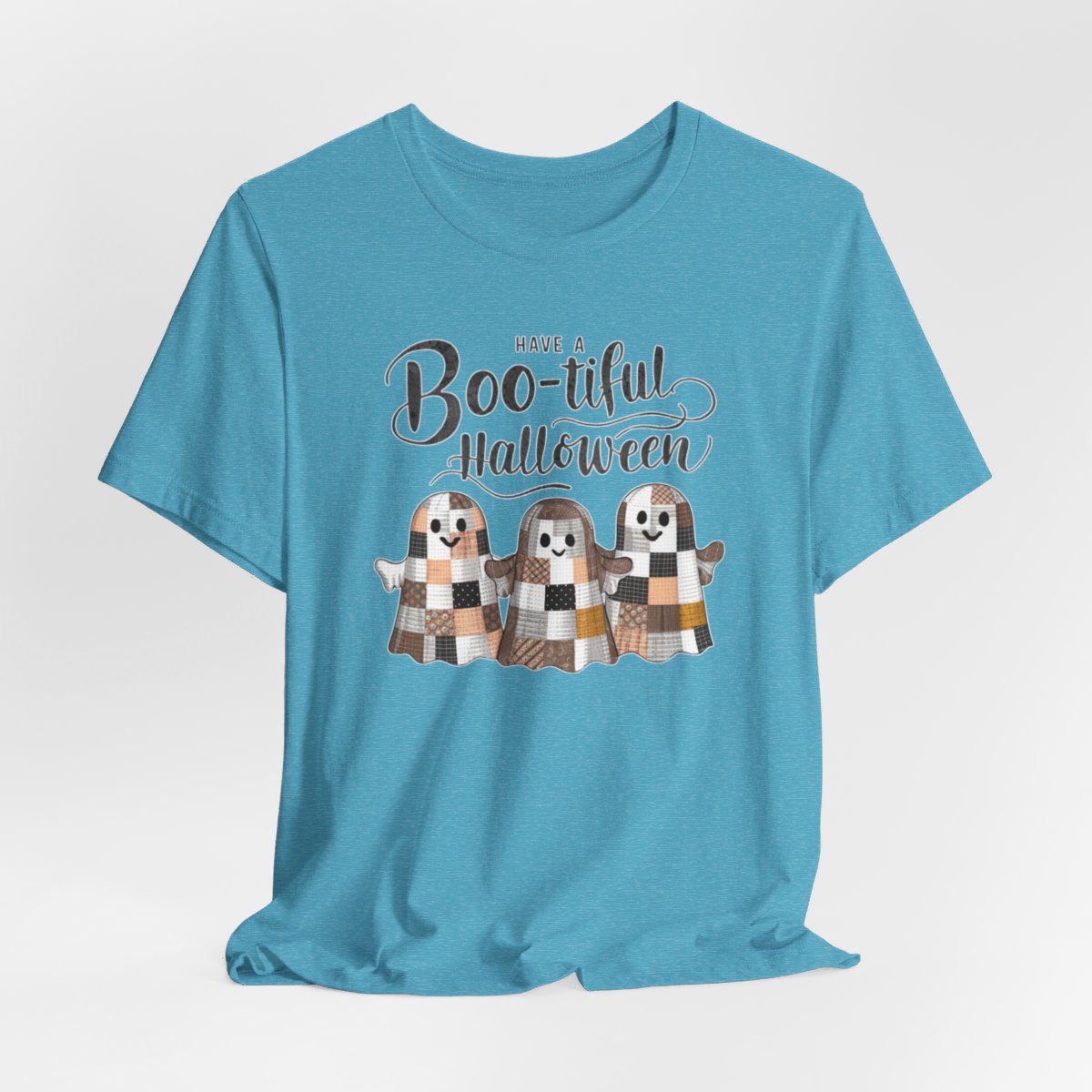 A Heather Aqua Halloween Quilting T-Shirt featuring three smiling patchwork ghosts made of various quilt patterns in autumn colors. Text reads "Have a Boo-tiful Halloween" in stylized lettering. Perfect for quilters who love Halloween.