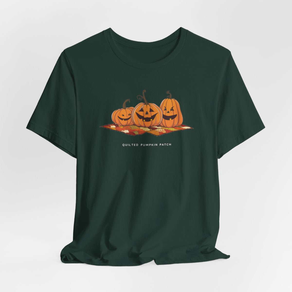 A Forest T-shirt with three smiling jack-o'-lanterns sitting on a colorful autumn-themed quilt. The pumpkins have carved faces with various expressions, and the quilt beneath them features a patchwork of fall colors. Text below reads "QUILTED PUMPKIN PATCH".