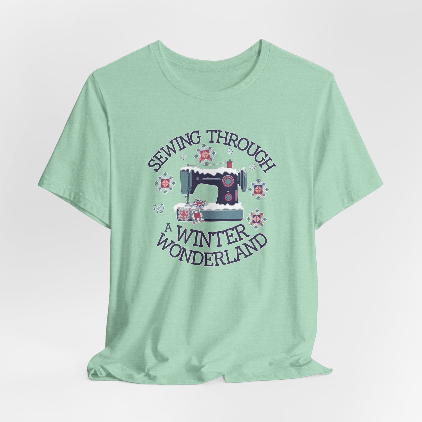 A Heather Mint winter quilting t-shirt featuring  a sewing machine covered in snow, surrounded by quilted snowflakes, with the phrase "Sewing Through a Winter Wonderland" above and below the image.