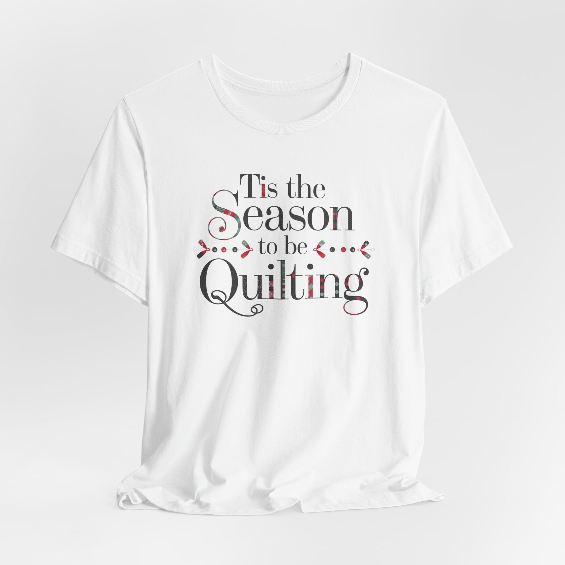 A White festive t-shirt featuring the phrase "'Tis the Season to be Quilting," with playful sewing-inspired elements like needles, buttons, and fabric patterns integrated into the typography.