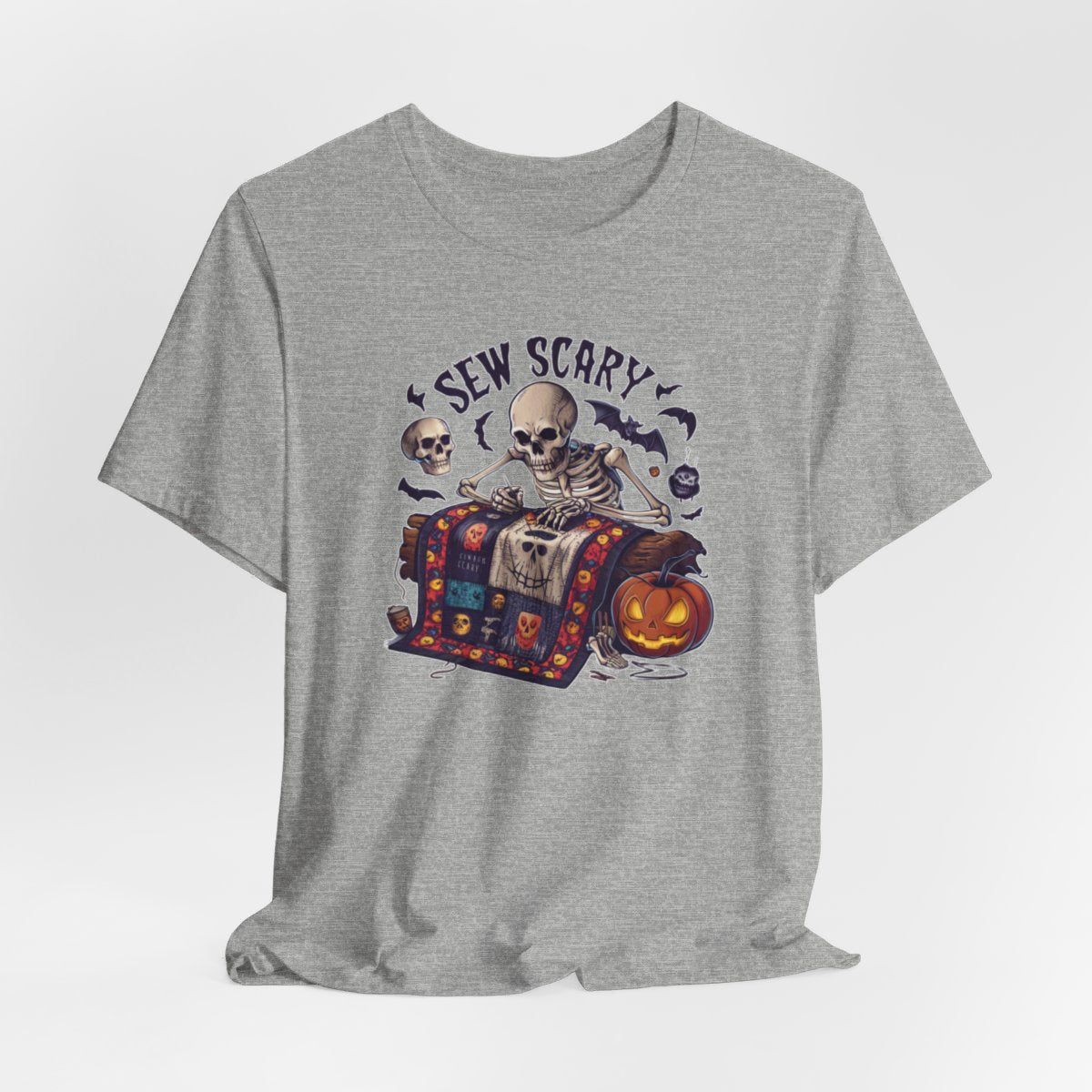An Athletic Heather Halloween Quilting T-Shirt featuring a skeleton sewing Halloween quilt. Colorful patchwork with spooky motifs. Bats and pumpkins surround skeleton. Text reads "Sew Scary". Ideal for quilters who love Halloween.