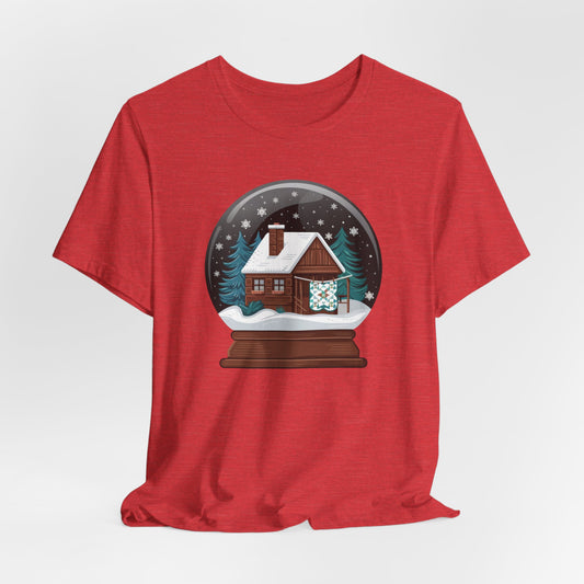 A Heather Red christmas quilting t-shirt featuring a cozy log cabin covered in snow, with a quilt hanging outside, set inside a snow globe with swirling snowflakes, blending quilting and holiday warmth.