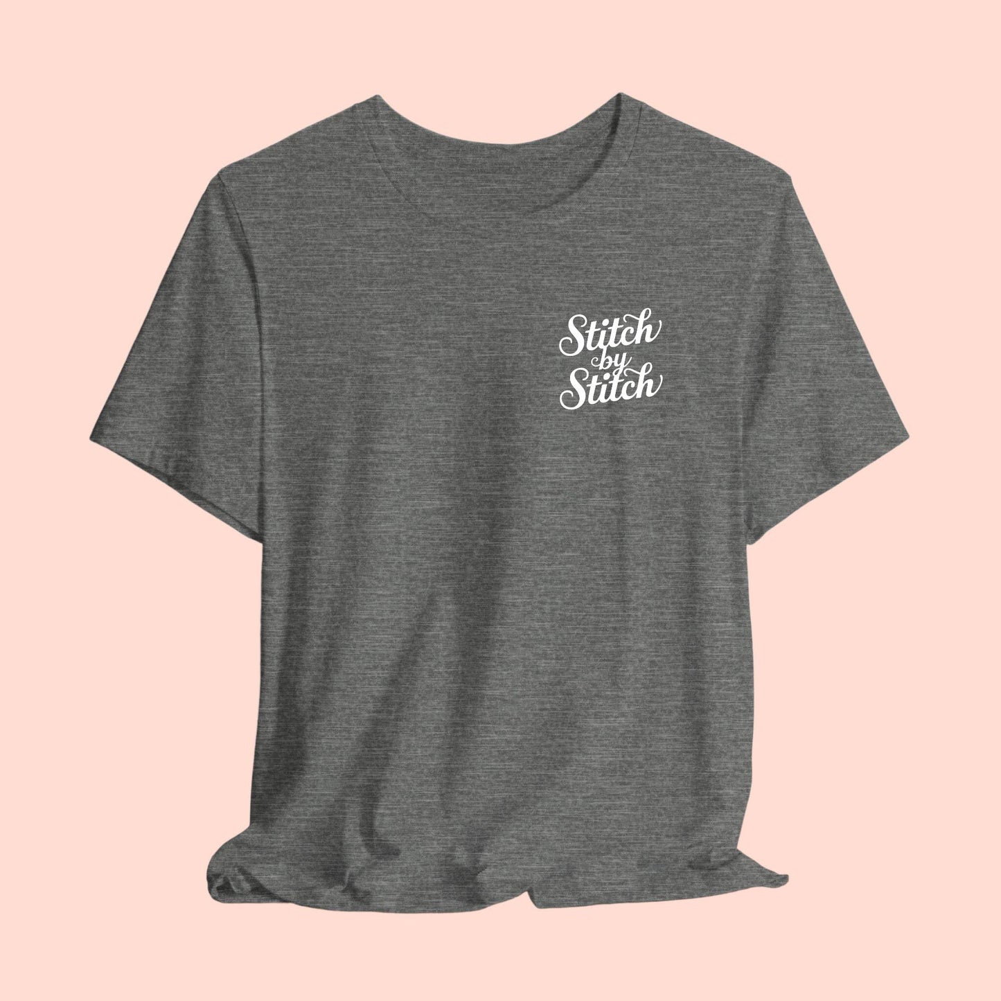 A T-Shirt featuring the phrase "Stitch by Stitch". The word "Stitch" is written twice in a flowing script font, while "by" is centered between them in a smaller, sans-serif font. The design has a clean, minimalist aesthetic.