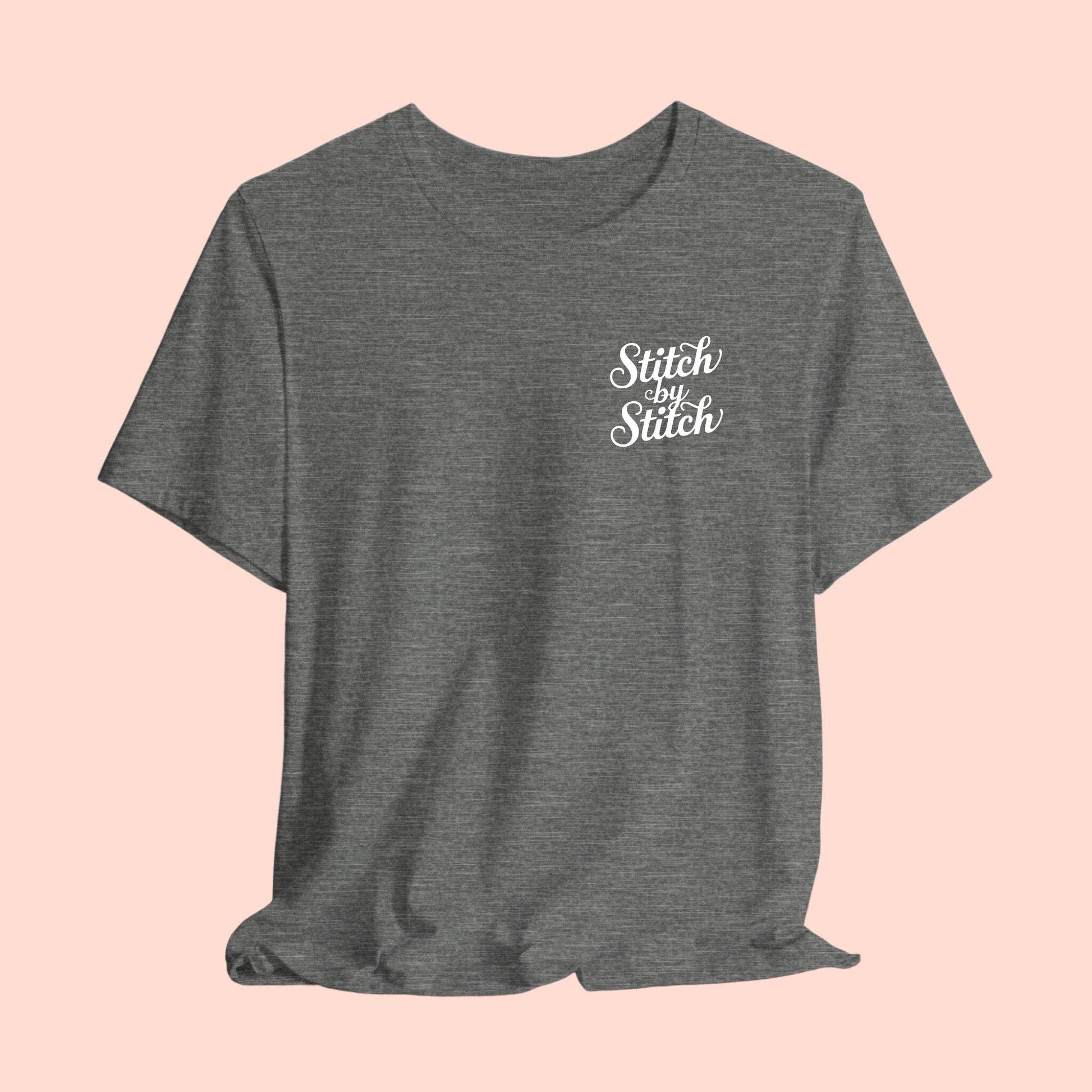 A T-Shirt featuring the phrase "Stitch by Stitch". The word "Stitch" is written twice in a flowing script font, while "by" is centered between them in a smaller, sans-serif font. The design has a clean, minimalist aesthetic.