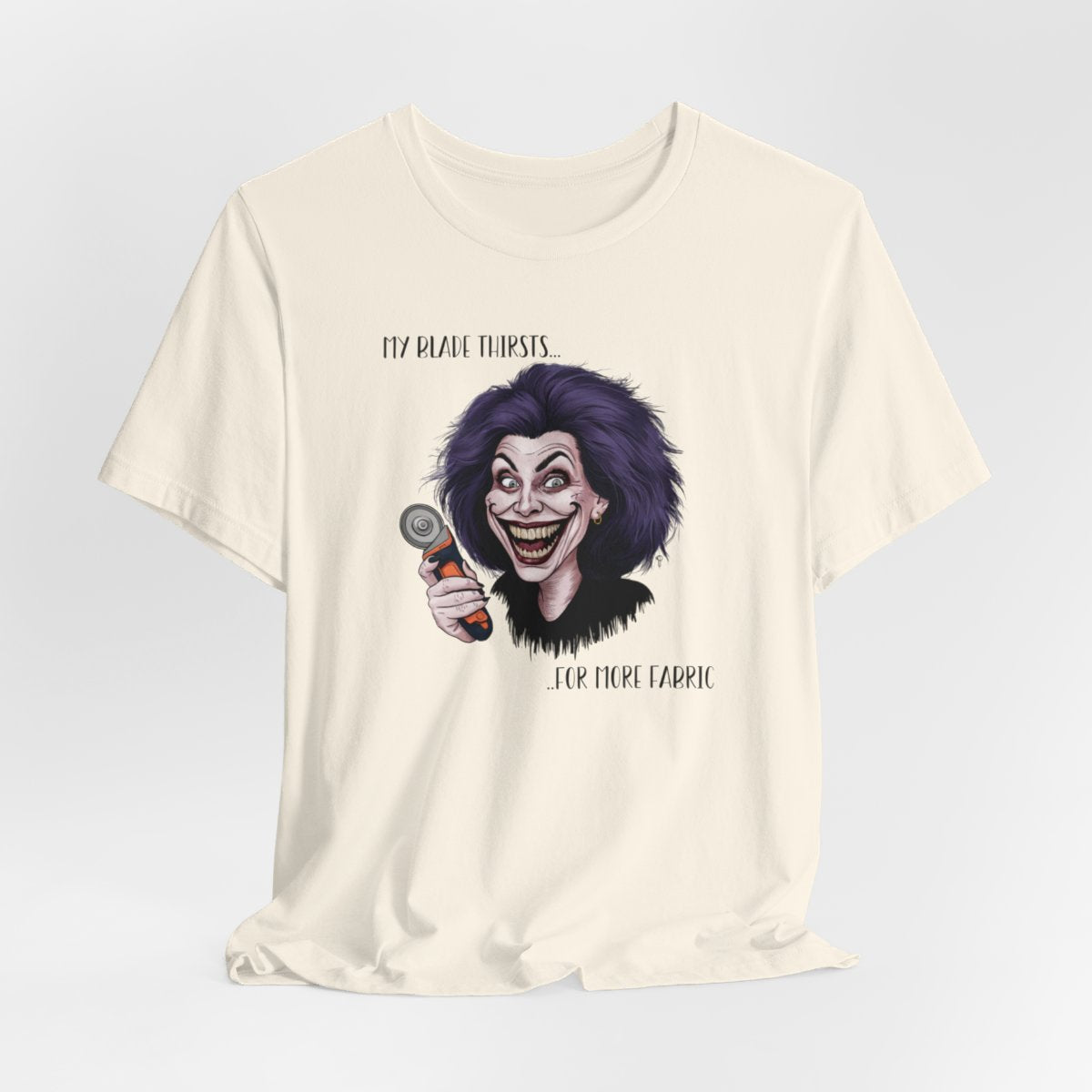 A Natural T-shirt showing a cartoonish character with wild purple hair and an exaggerated grin, holding a rotary cutter. Text reads "My blade thirsts... for more fabric", humorously depicting a quilter's endless desire for new materials.