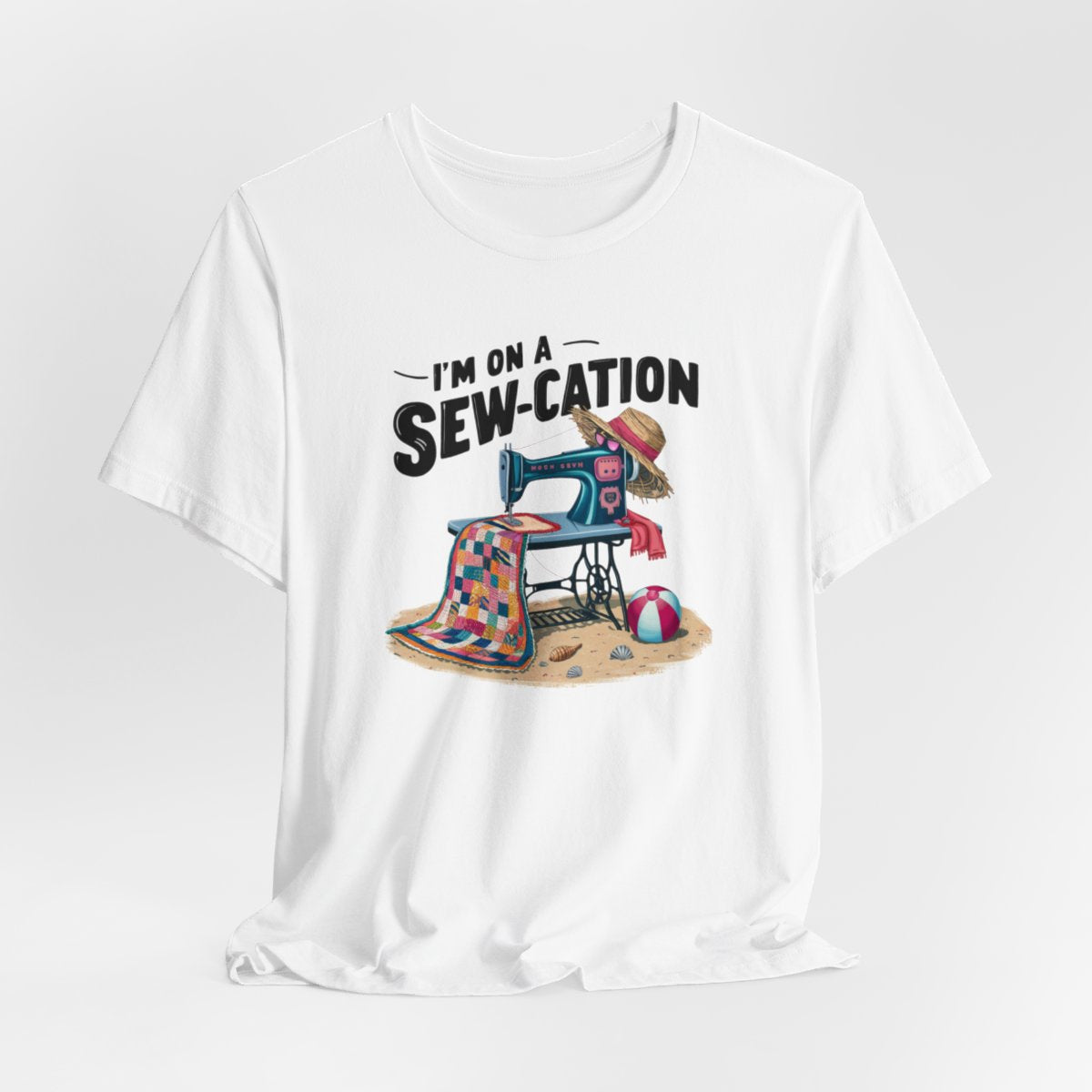 A White fun Quilting T-shirt of a vintage sewing machine on a beach with a colorful quilt, sun hat, and beach ball. Text above reads "I'm on a Sew-cation" in bold letters.