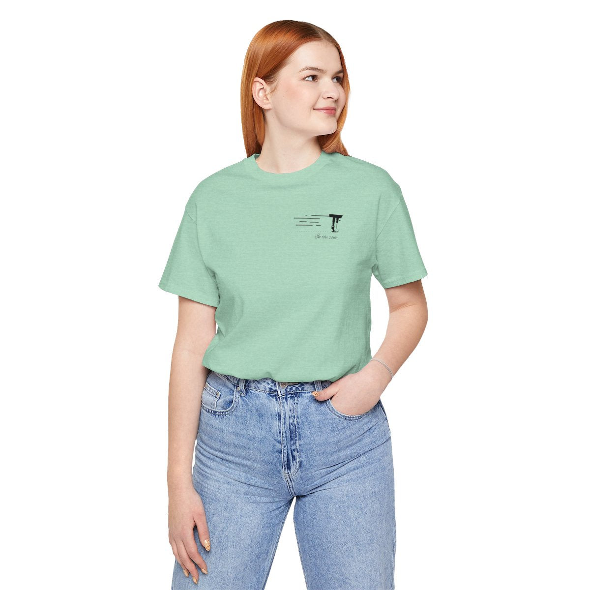 A Heather Mint T-shirt featuring a minimalist black silhouette of a sewing machine arm with motion lines. Below, the phrase "In the zone" is written in cursive script. The design conveys the focus and rhythm of quilting.