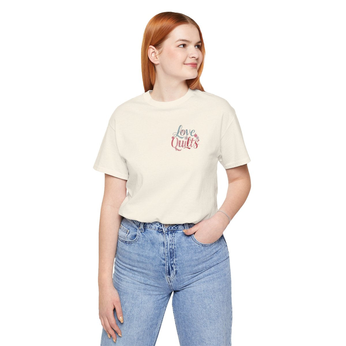 A Natural t-shirt spelling out "Love Quilts". The word "Love" is in teal, while "Quilts" features various shades of pink and red. Small flowers in white and pink adorn the letters, creating a floral and quilting-inspired artwork.
