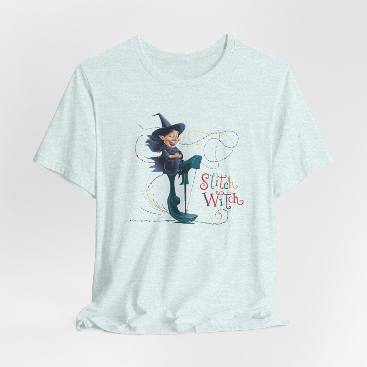 A Heather Ice Blue T-shirt with a cartoon illustration of a smiling witch with pointed hat riding a teal sewing machine. Colorful stitches swirl around her in the air. Text reads "Stitch Witch" in playful, multicolored letters.