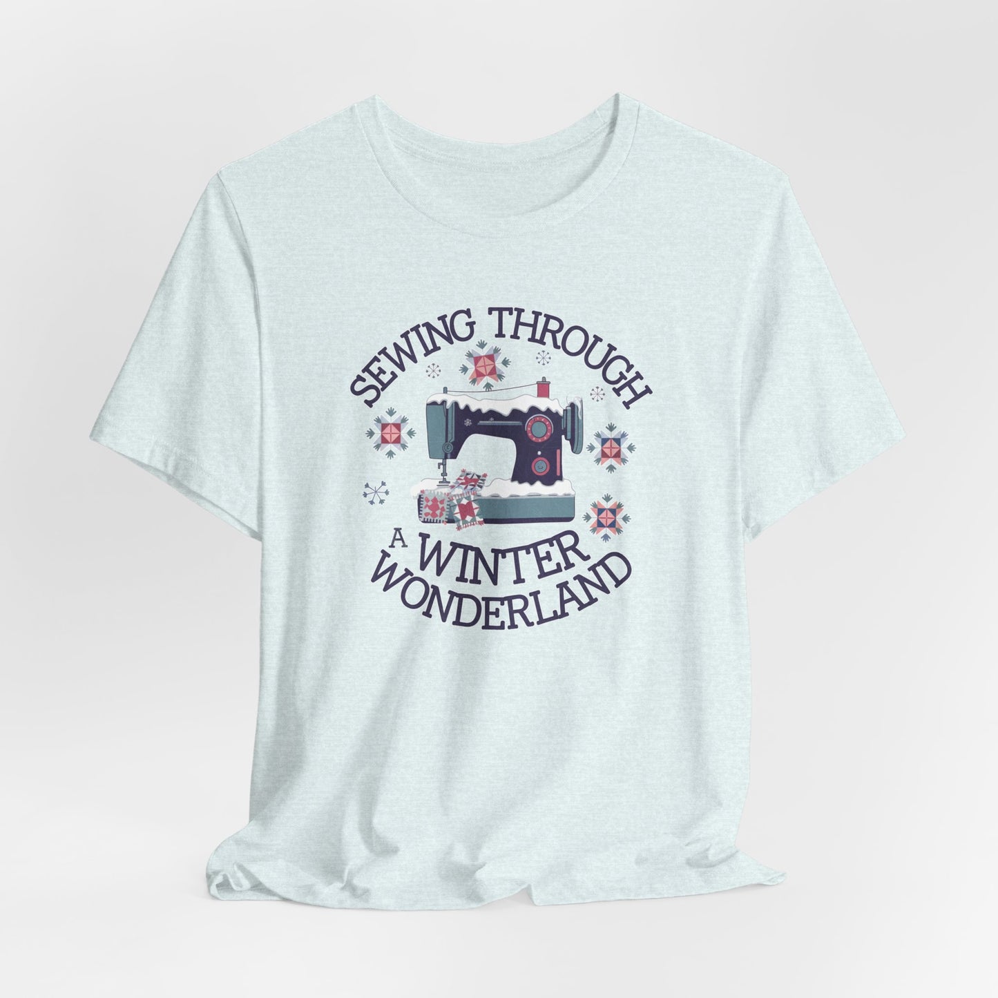 A Heather Ice Blue winter quilting t-shirt featuring  a sewing machine covered in snow, surrounded by quilted snowflakes, with the phrase "Sewing Through a Winter Wonderland" above and below the image.
