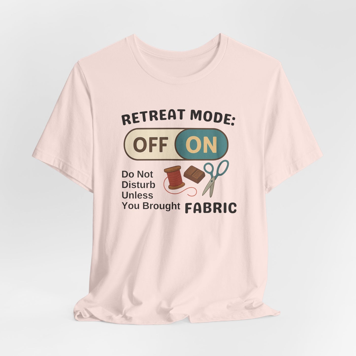 A A Soft Pink t-shirt with a toggle switch that says "Retreat Mode: OFF/ON" surrounded by a spool of thread, chocolate, and scissors. Text below reads "Do not disturb unless you brought fabric." Fun gift for quilting retreats.