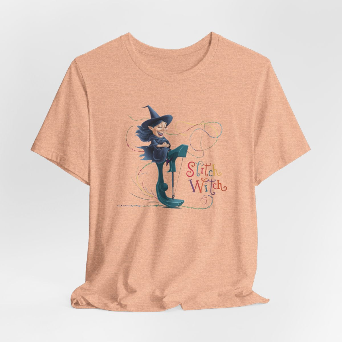 A Heather Peach T-shirt with a cartoon illustration of a smiling witch with pointed hat riding a teal sewing machine. Colorful stitches swirl around her in the air. Text reads "Stitch Witch" in playful, multicolored letters.