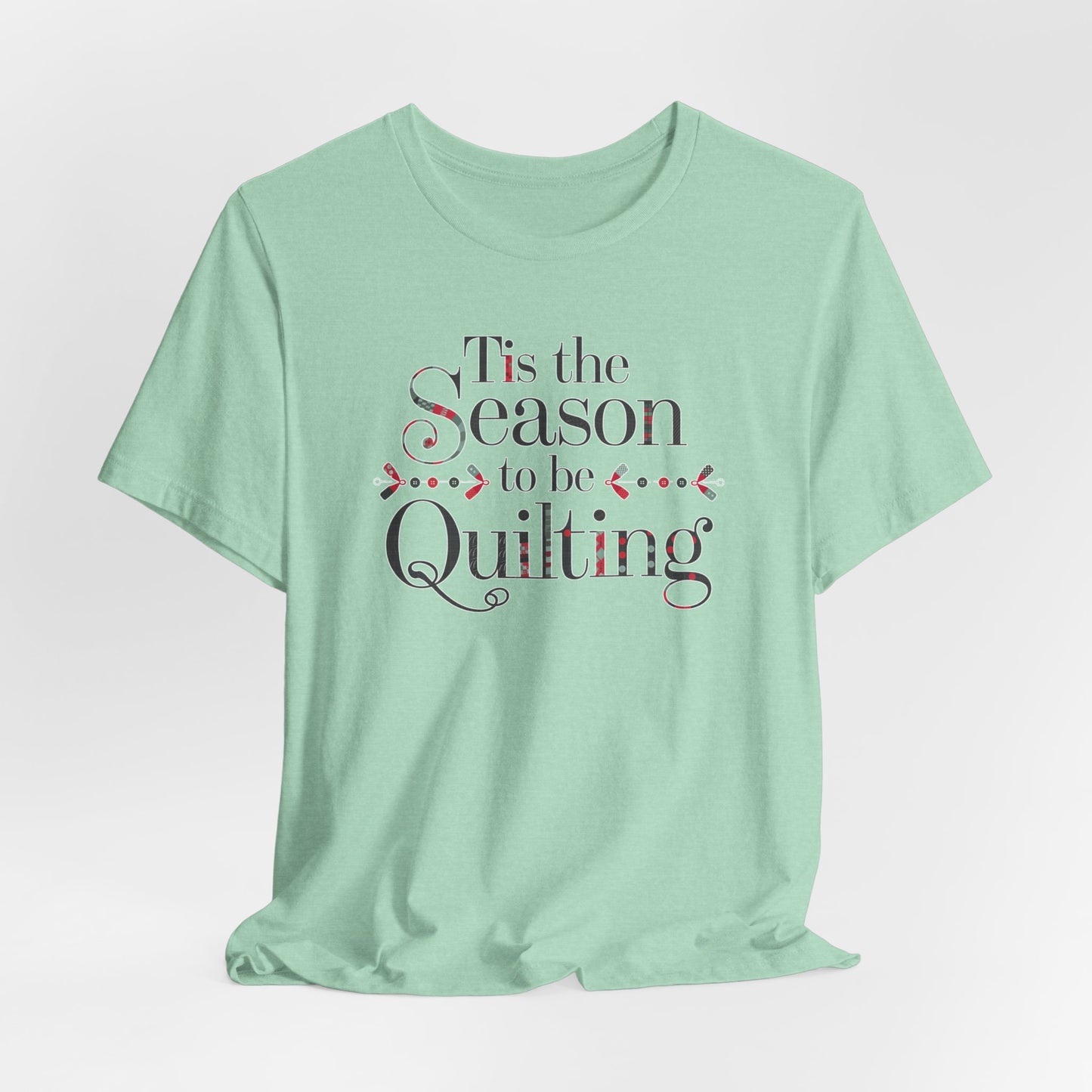A Heather Mint festive t-shirt featuring the phrase "'Tis the Season to be Quilting," with playful sewing-inspired elements like needles, buttons, and fabric patterns integrated into the typography.