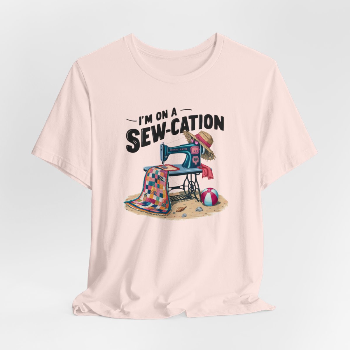 A Soft Pink fun Quilting T-shirt of a vintage sewing machine on a beach with a colorful quilt, sun hat, and beach ball. Text above reads "I'm on a Sew-cation" in bold letters.
