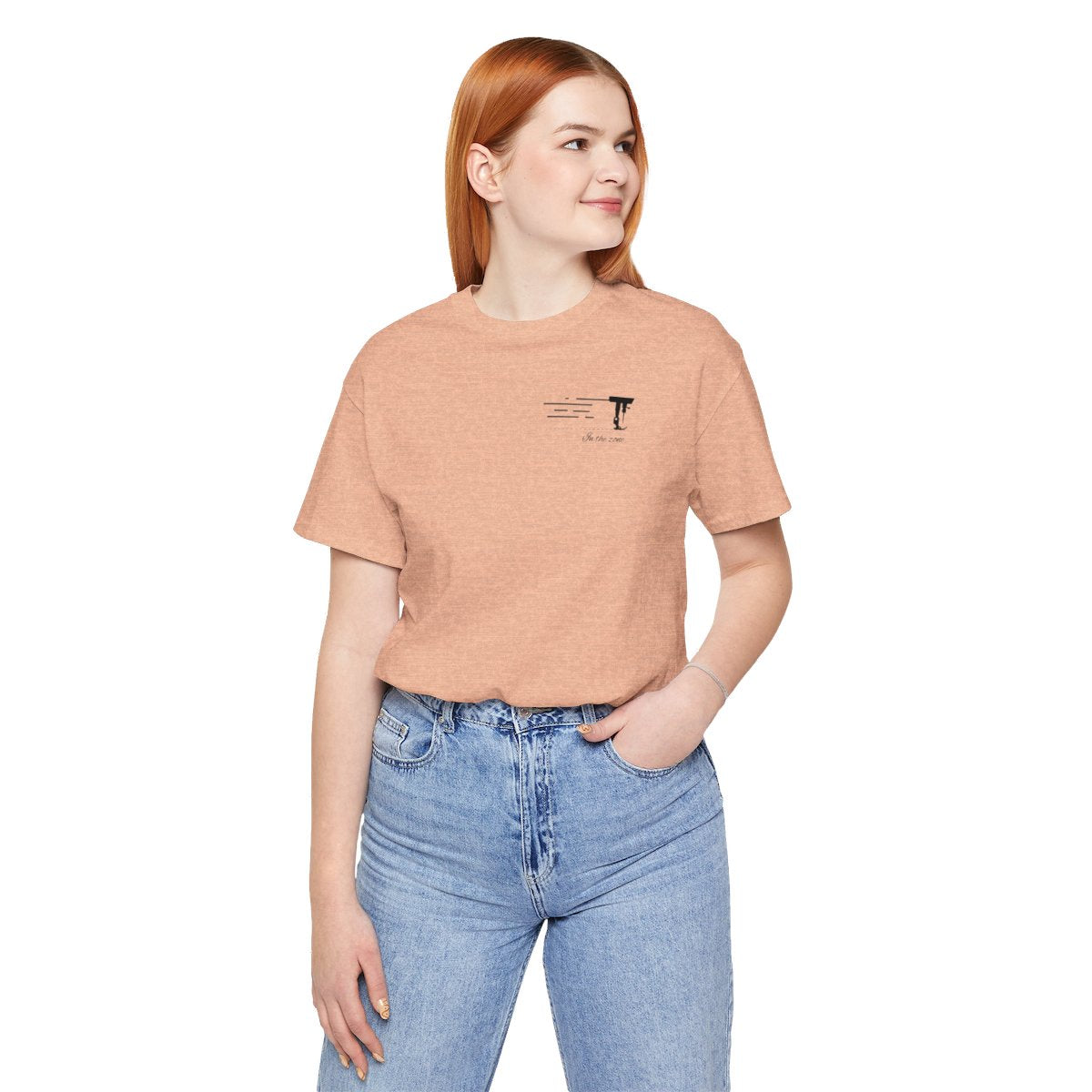 A Heather Peach T-shirt featuring a minimalist black silhouette of a sewing machine arm with motion lines. Below, the phrase "In the zone" is written in cursive script. The design conveys the focus and rhythm of quilting.