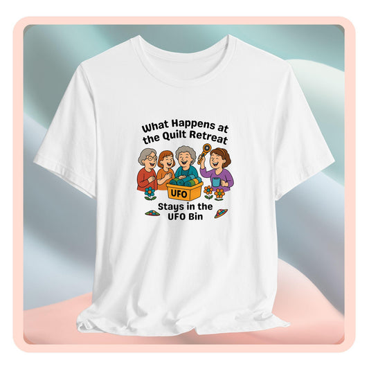 A A t-shirt featuring a cartoon design of four laughing women at a quilt retreat with a bin labeled “UFO” full of fabric. Surrounded by patchwork flying saucers and flowers. Text reads “What happens at the quilt retreat stays in the UFO bin.”