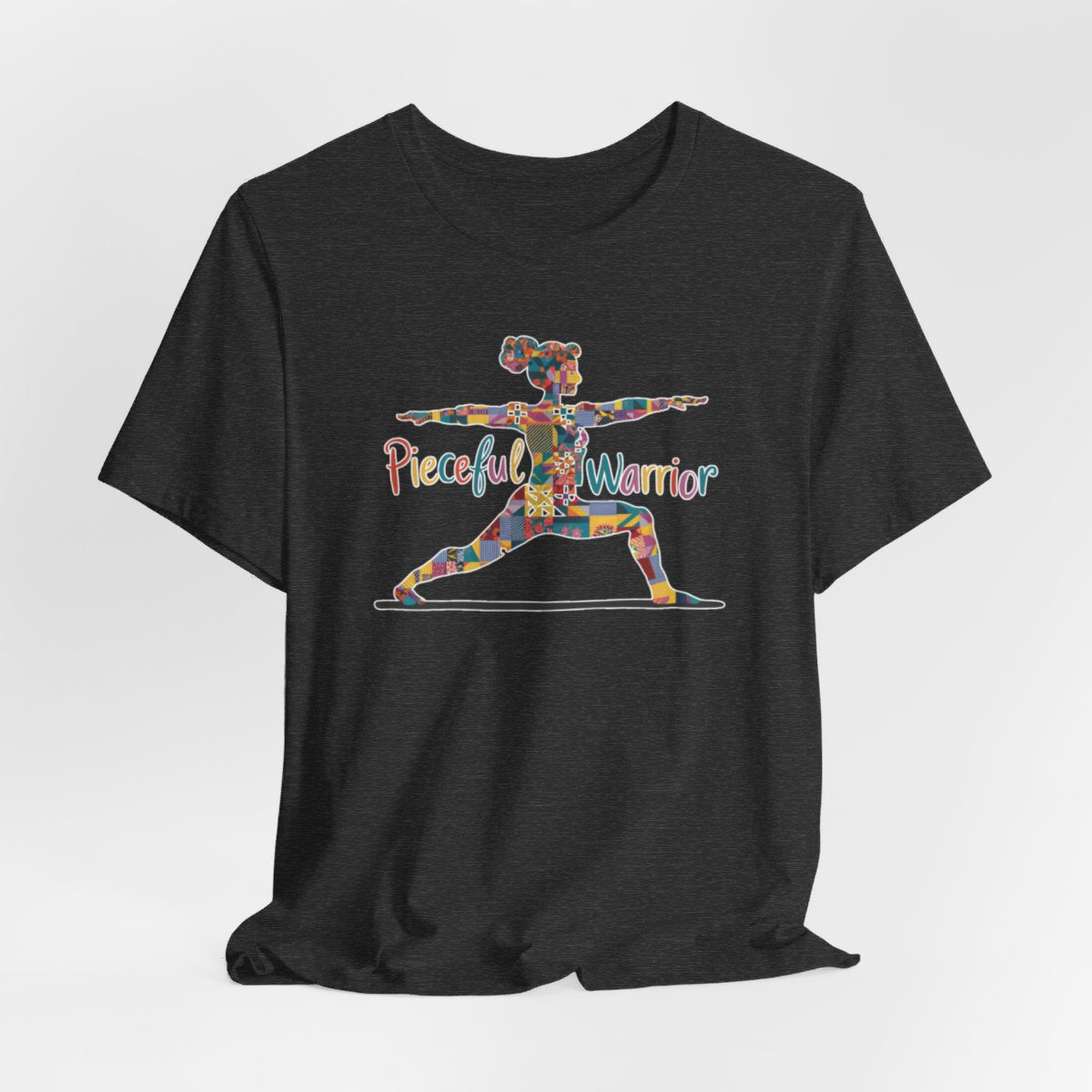 A Dark Grey Heather Tee featuring a colorful patchwork silhouette of woman in yoga warrior pose. Text reads "Pieceful Warrior" in multicolored lettering.