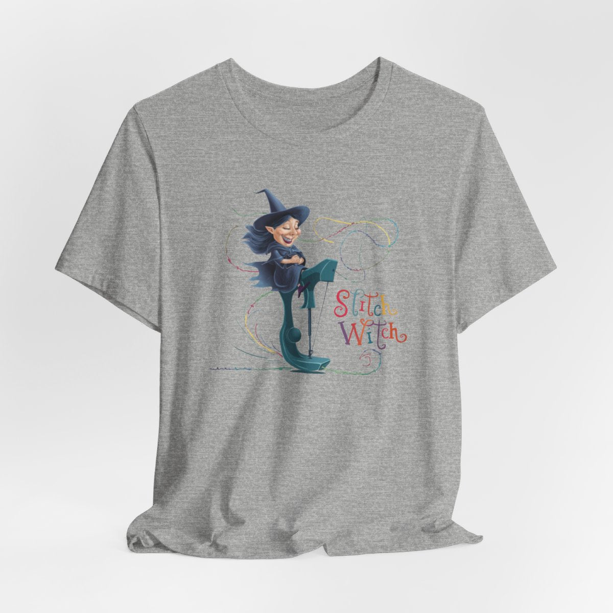 An Athletic Heather T-shirt with a cartoon illustration of a smiling witch with pointed hat riding a teal sewing machine. Colorful stitches swirl around her in the air. Text reads "Stitch Witch" in playful, multicolored letters.