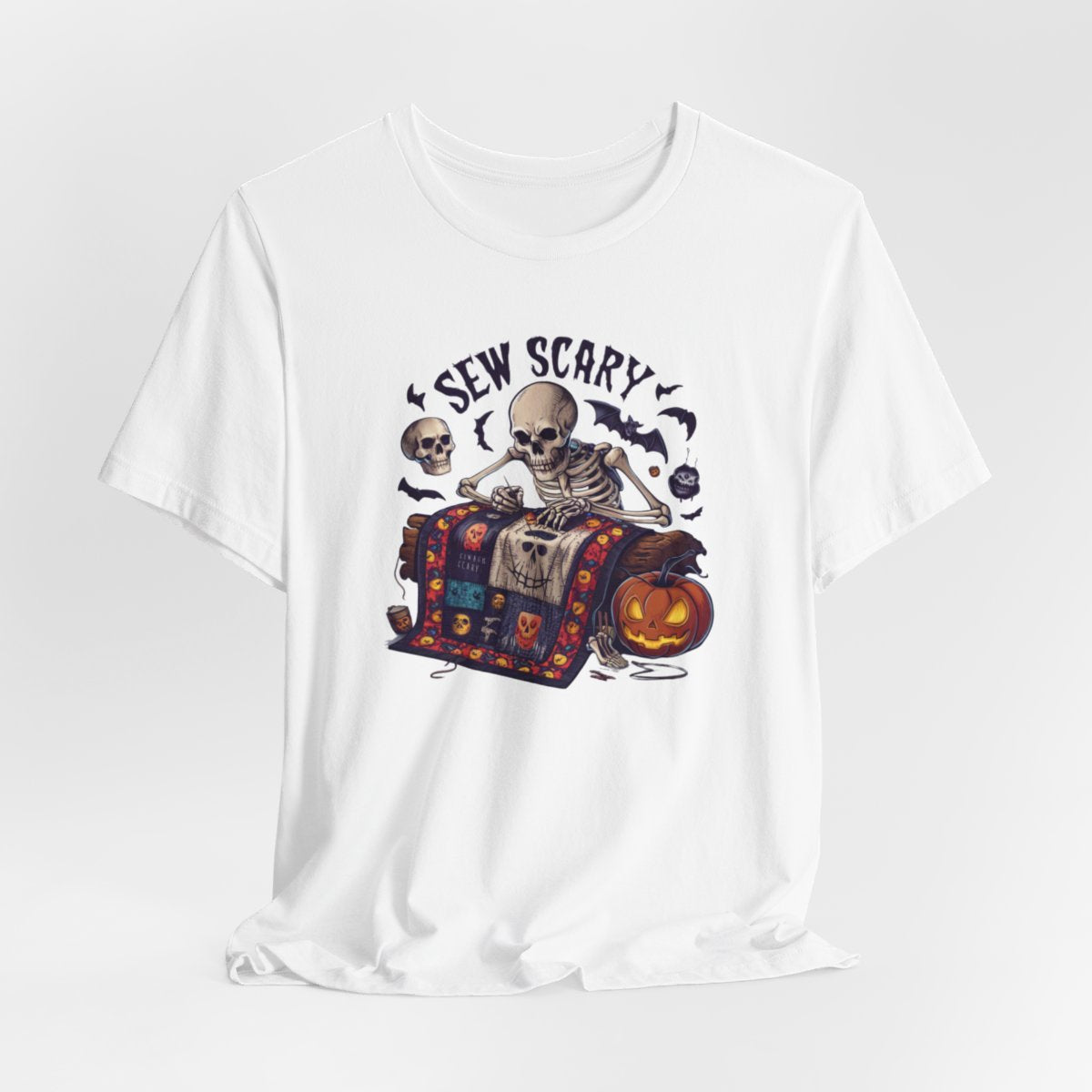 A White Halloween Quilting T-Shirt featuring a skeleton sewing Halloween quilt. Colorful patchwork with spooky motifs. Bats and pumpkins surround skeleton. Text reads "Sew Scary". Ideal for quilters who love Halloween.