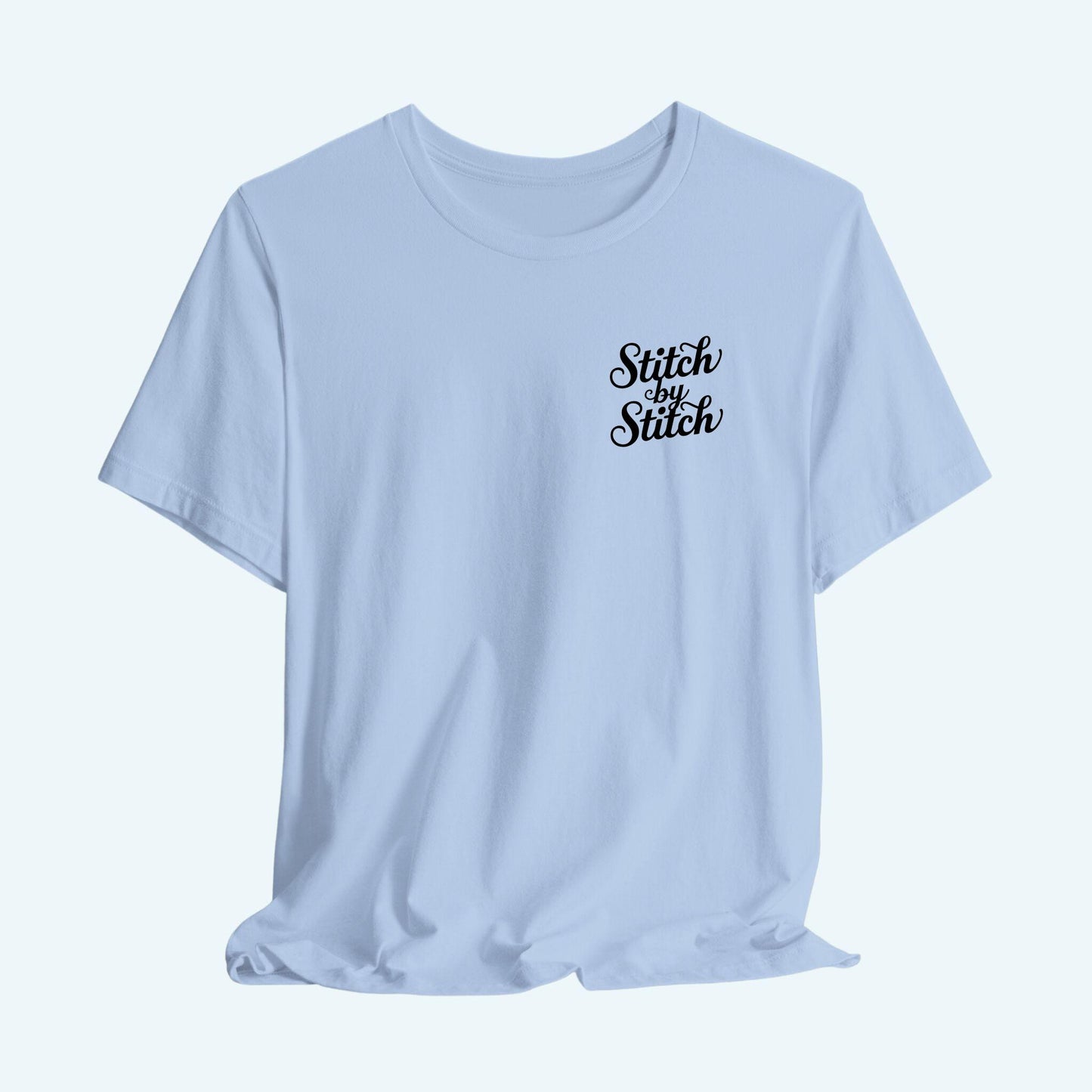 A T-Shirt featuring the phrase "Stitch by Stitch". The word "Stitch" is written twice in a flowing script font, while "by" is centered between them in a smaller, sans-serif font. The design has a clean, minimalist aesthetic.