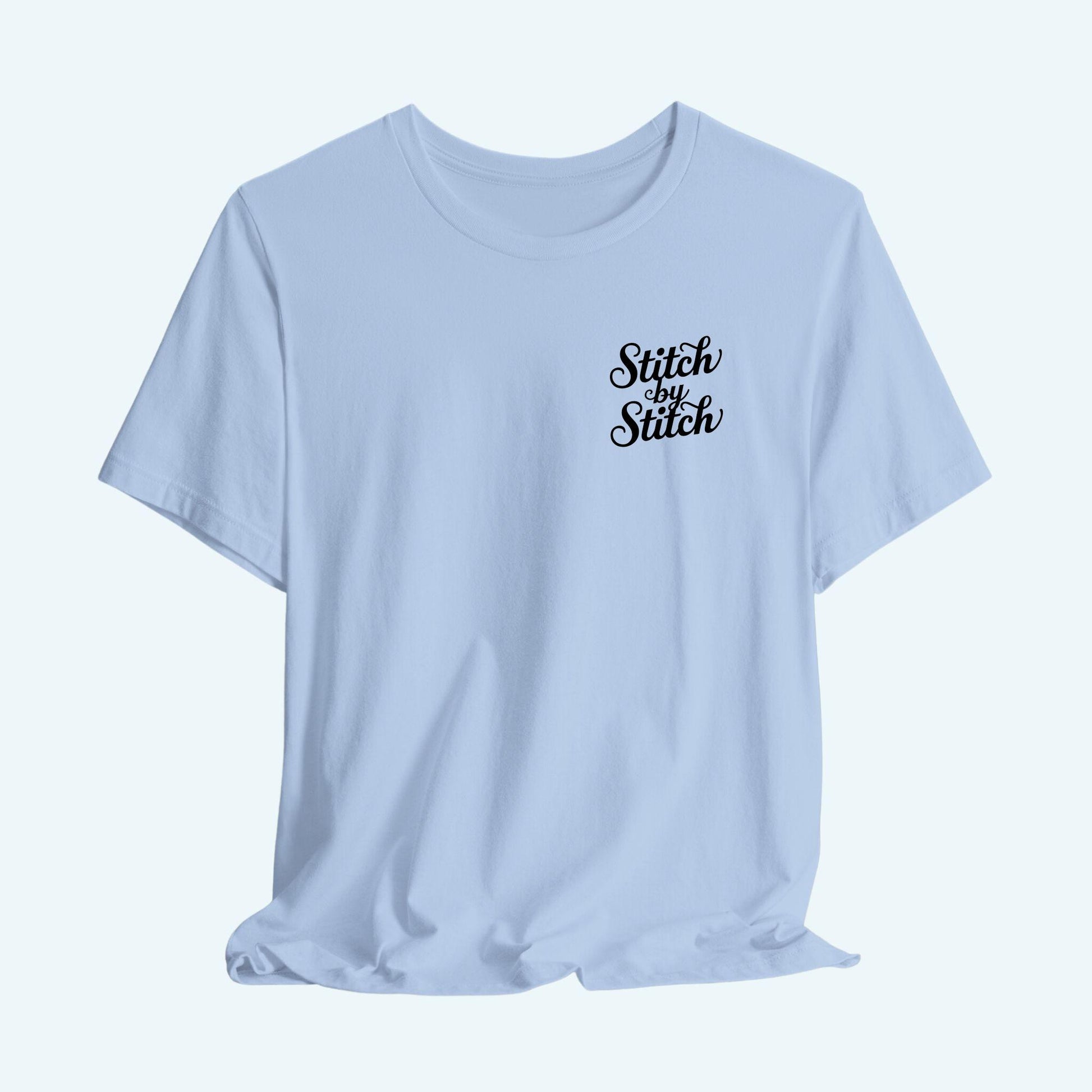 A T-Shirt featuring the phrase "Stitch by Stitch". The word "Stitch" is written twice in a flowing script font, while "by" is centered between them in a smaller, sans-serif font. The design has a clean, minimalist aesthetic.