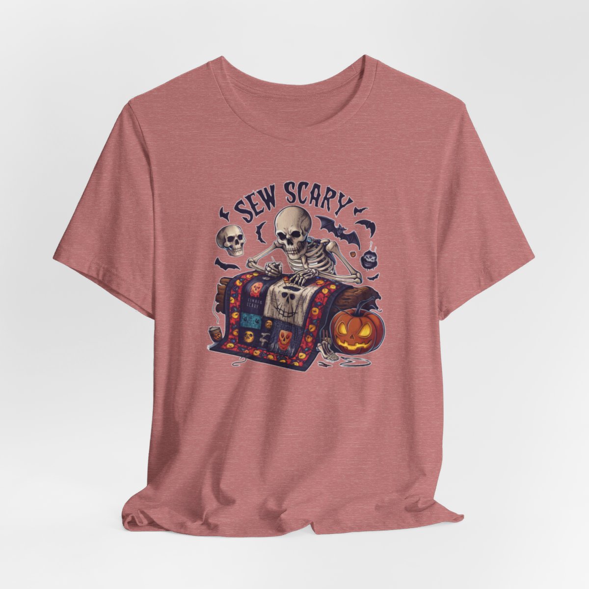 A Heather Mauve Halloween Quilting T-Shirt featuring a skeleton sewing Halloween quilt. Colorful patchwork with spooky motifs. Bats and pumpkins surround skeleton. Text reads "Sew Scary". Ideal for quilters who love Halloween.