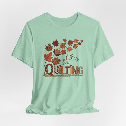 A Heather Mint thanksgiving  quilting t-shirt featuring autumn leaves with various quilting patterns inside. Text reads "falling for QUILTING" with letters in patchwork style. Warm fall colors and quilting motifs combine in a seasonal design.