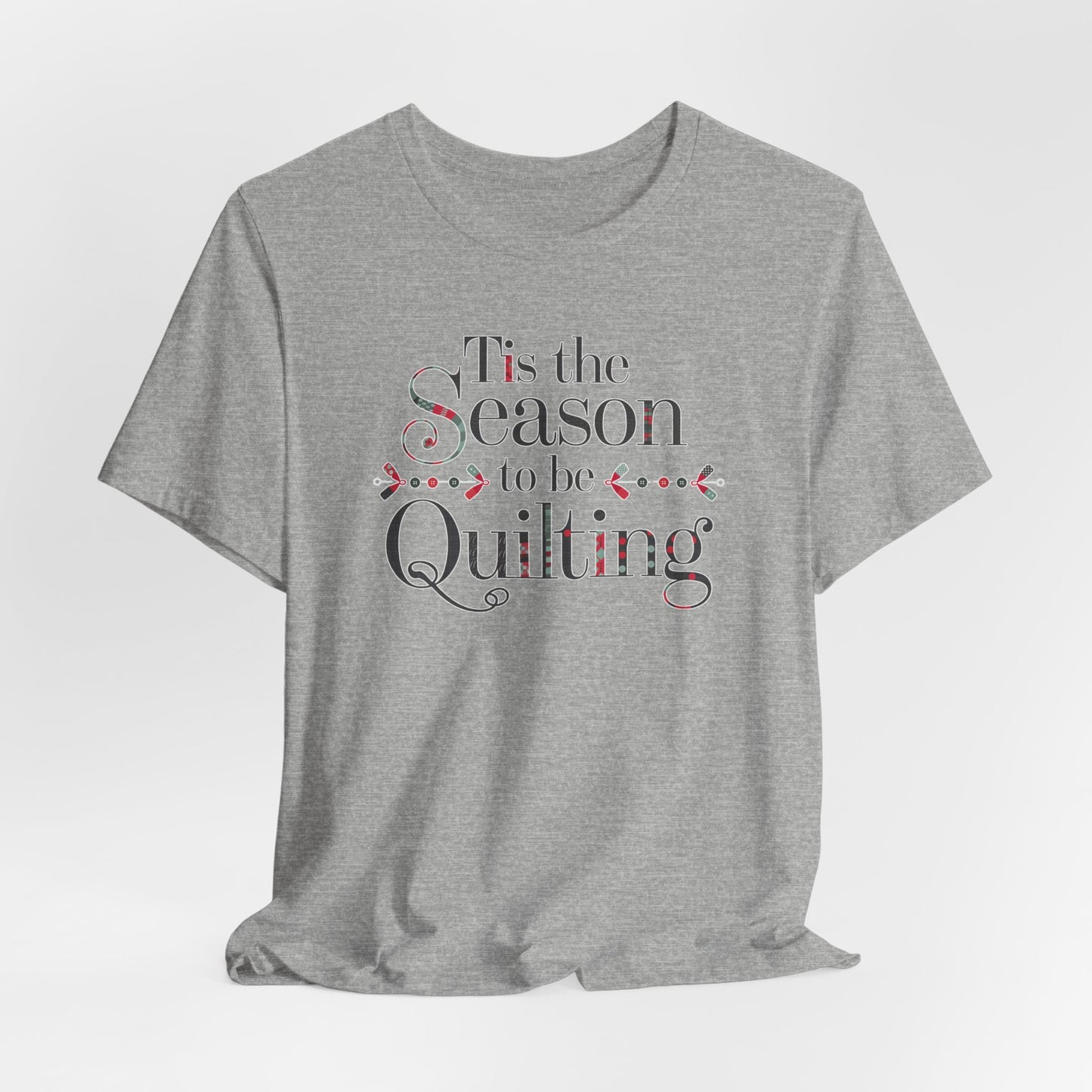 An Athletic Heather festive t-shirt featuring the phrase "'Tis the Season to be Quilting," with playful sewing-inspired elements like needles, buttons, and fabric patterns integrated into the typography.