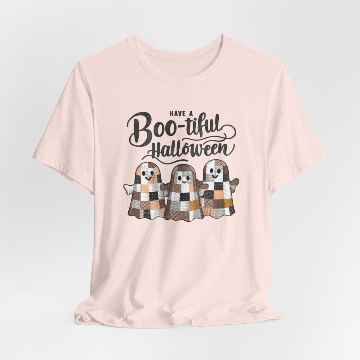 A Soft Pink Halloween Quilting T-Shirt featuring three smiling patchwork ghosts made of various quilt patterns in autumn colors. Text reads "Have a Boo-tiful Halloween" in stylized lettering. Perfect for quilters who love Halloween.