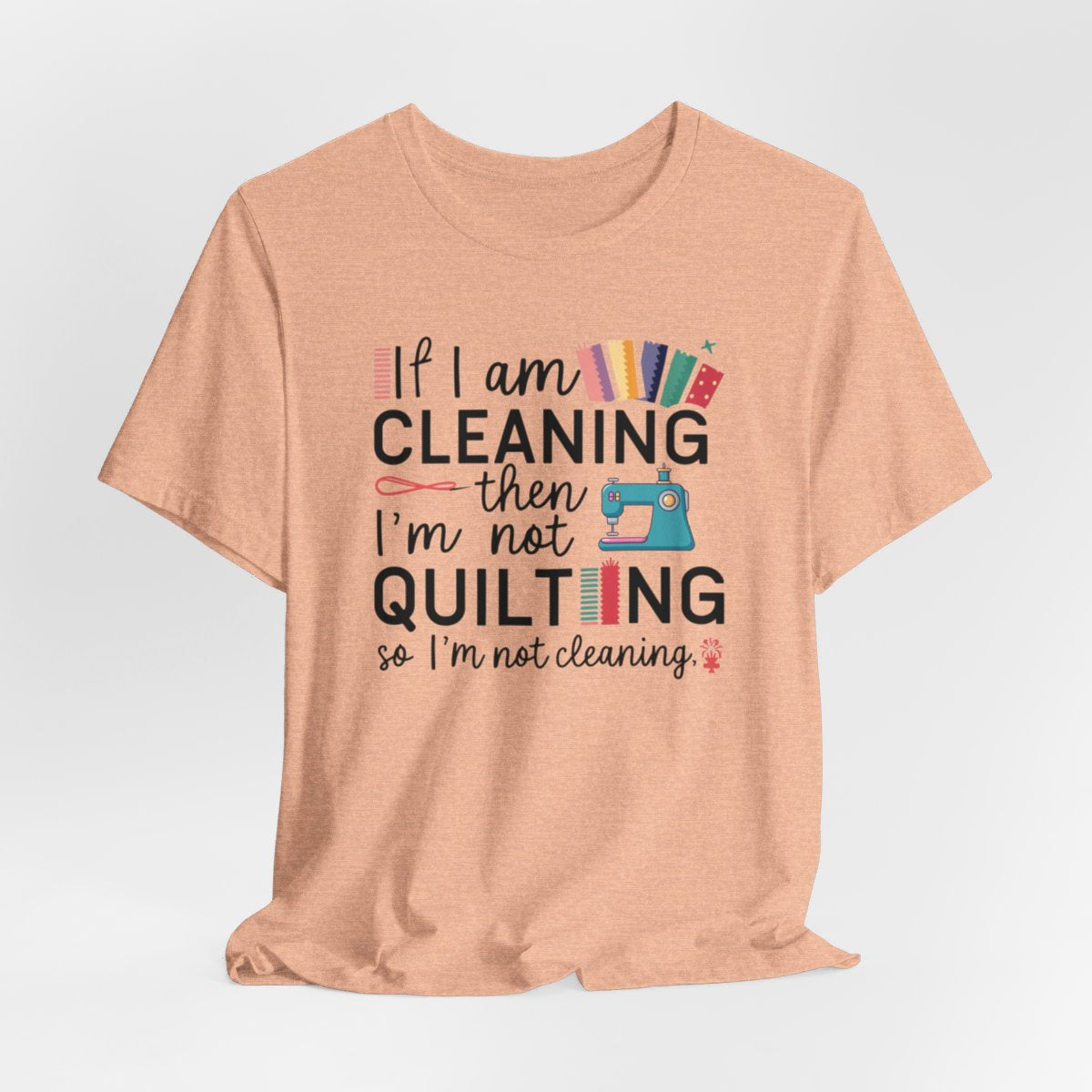 A Heather Peach funny quilting T-Shirt with the phrase If I am Cleaning then I'm not Quilting in a bold and cheerful font with images of quilting icons and supplies