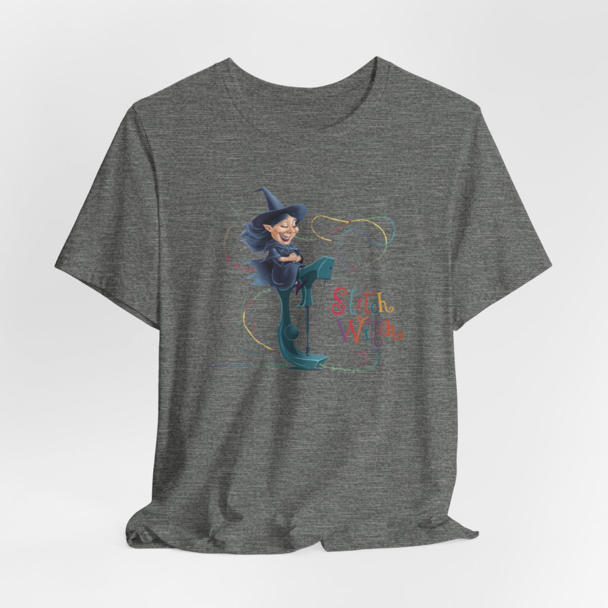 A Deep Heather T-shirt with a cartoon illustration of a smiling witch with pointed hat riding a teal sewing machine. Colorful stitches swirl around her in the air. Text reads "Stitch Witch" in playful, multicolored letters.