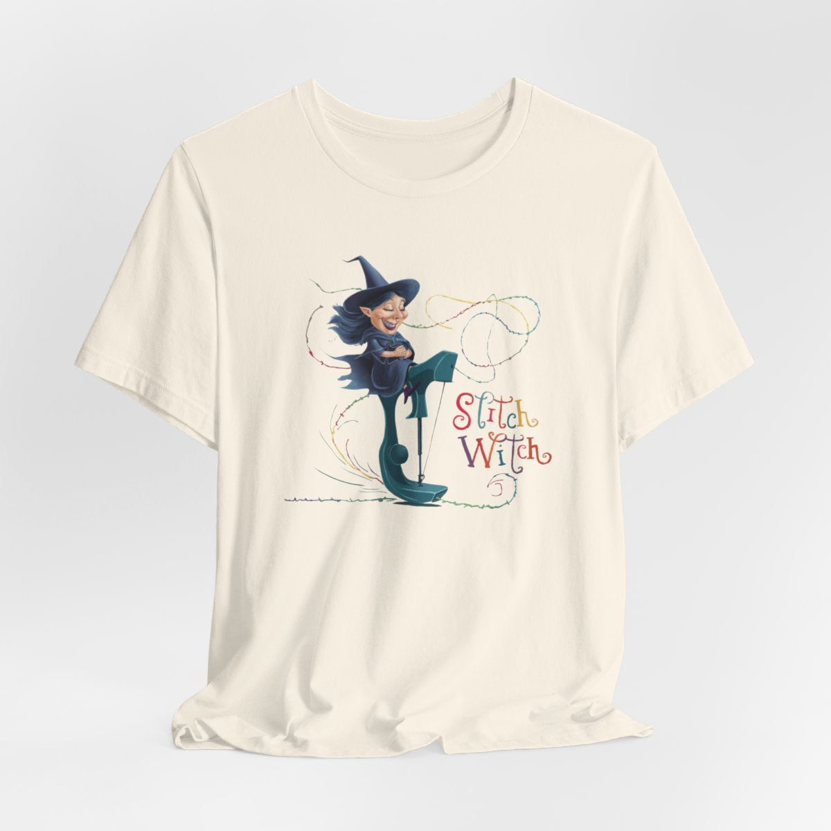 A Natural T-shirt with a cartoon illustration of a smiling witch with pointed hat riding a teal sewing machine. Colorful stitches swirl around her in the air. Text reads "Stitch Witch" in playful, multicolored letters.