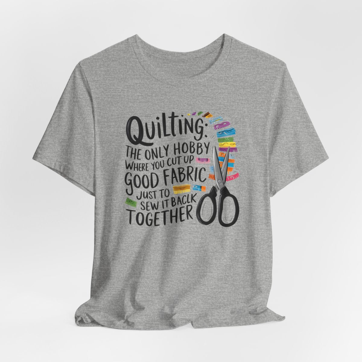 An Athletic Heather Quilting-themed T-Shirt with text "Quilting: The Only Hobby Where You Cut Up Good Fabric Just to Sew it Back Together" surrounded by colorful fabric scraps and scissors.